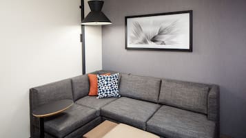 A gray sectional sofa with a small round table and a box on the floor. A black floor lamp and a framed abstract art piece are on the wall.