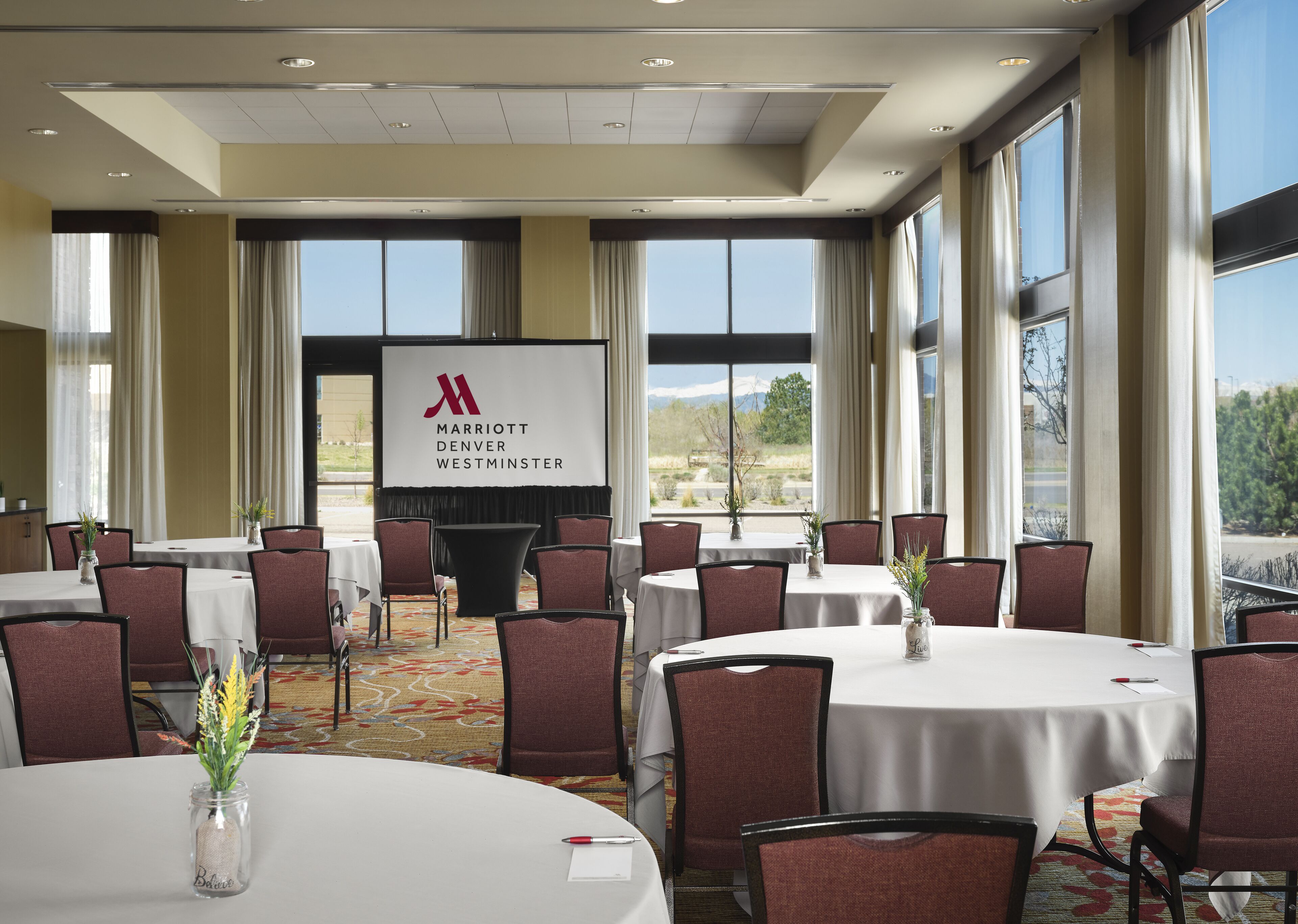 A Marriott Denver Westminster conference room with multiple tables and chairs, large windows, and a presentation screen.