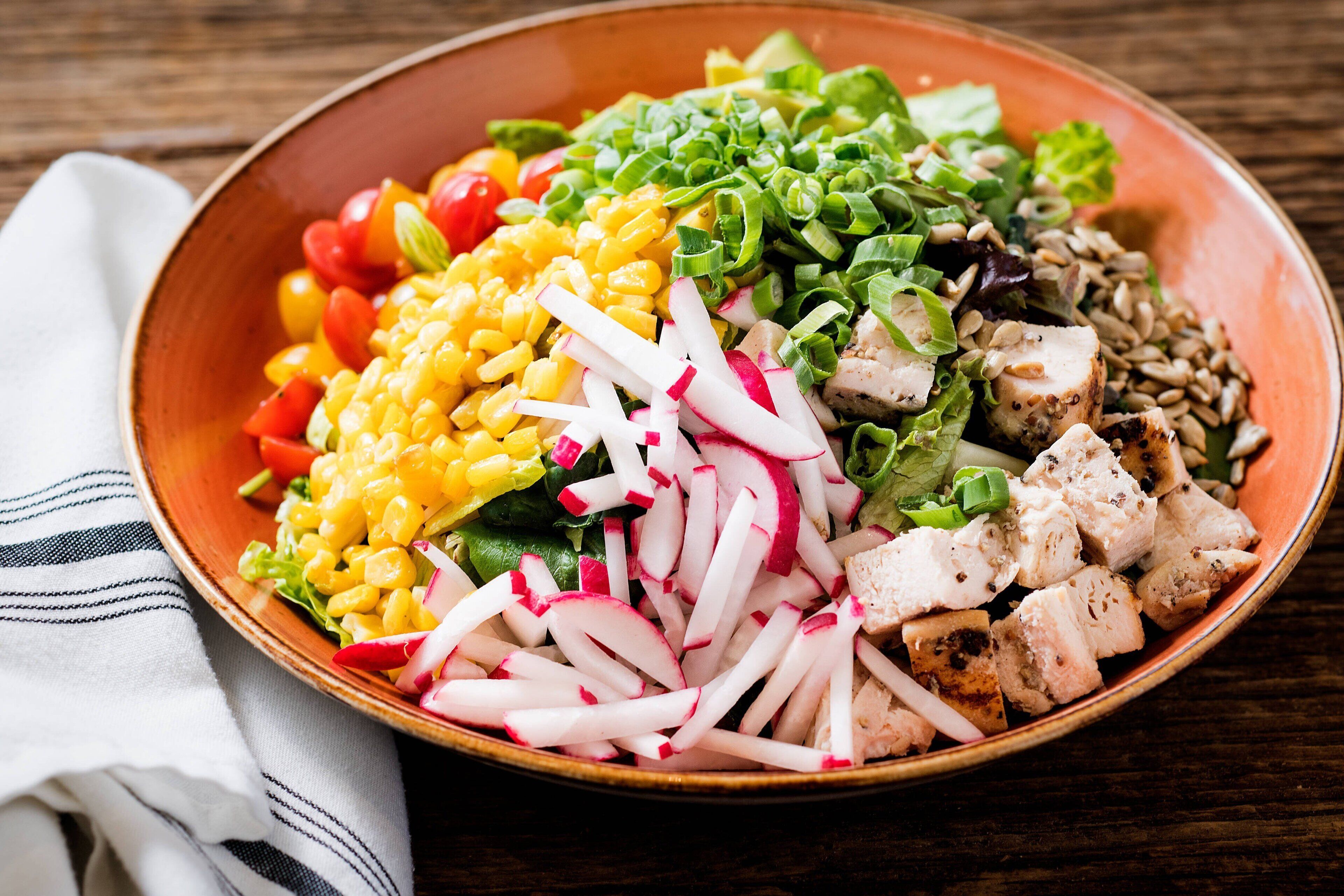 Un tazón colorido con ensalada, diversos vegetales y trozos de pollo sobre una mesa de madera.