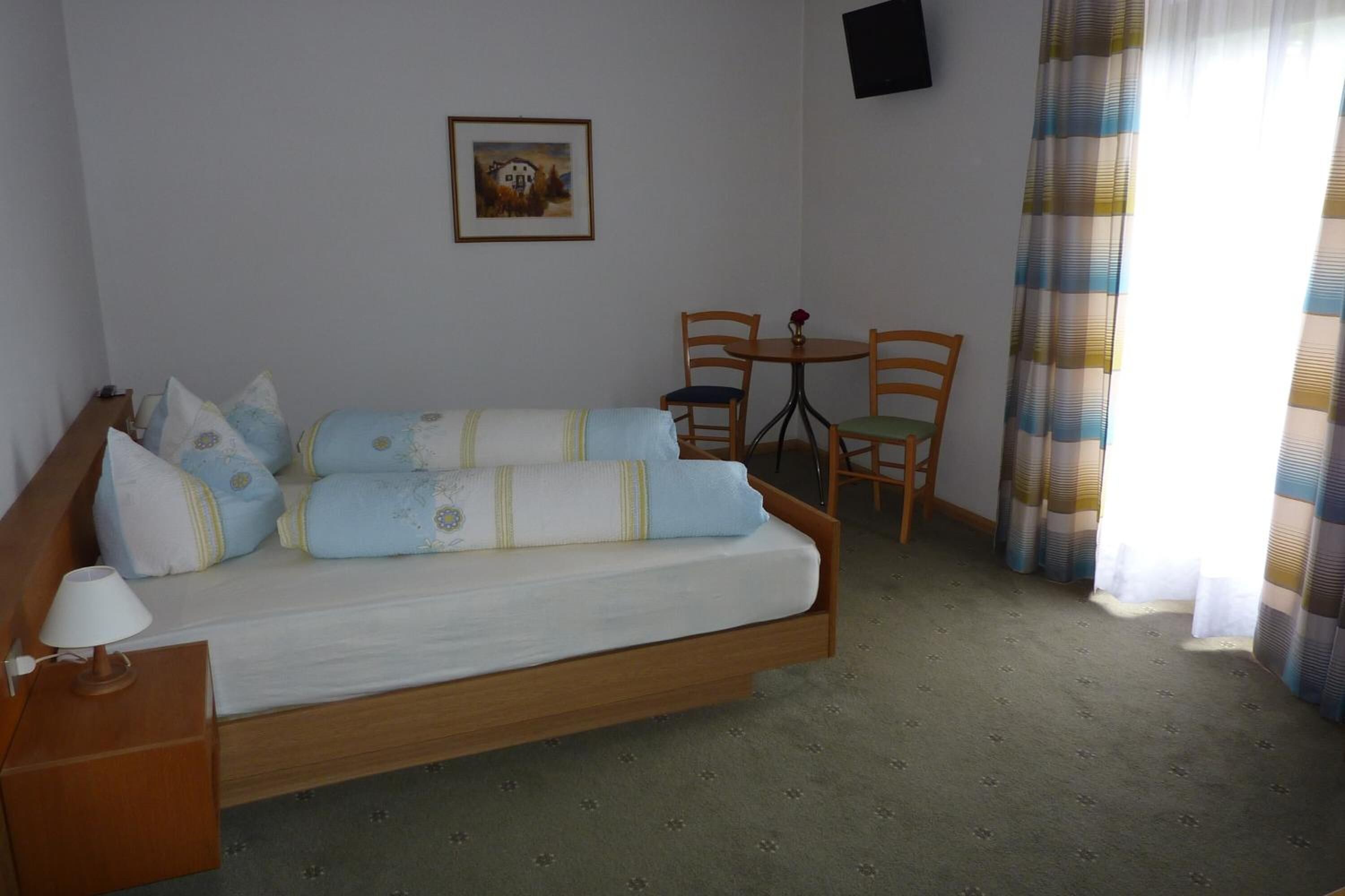 Una habitación con cama, una mesita con sillas, un cuadro en la pared y una ventana con cortinas.