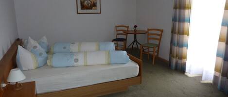 Una habitación con cama, una mesita con sillas, un cuadro en la pared y una ventana con cortinas.
