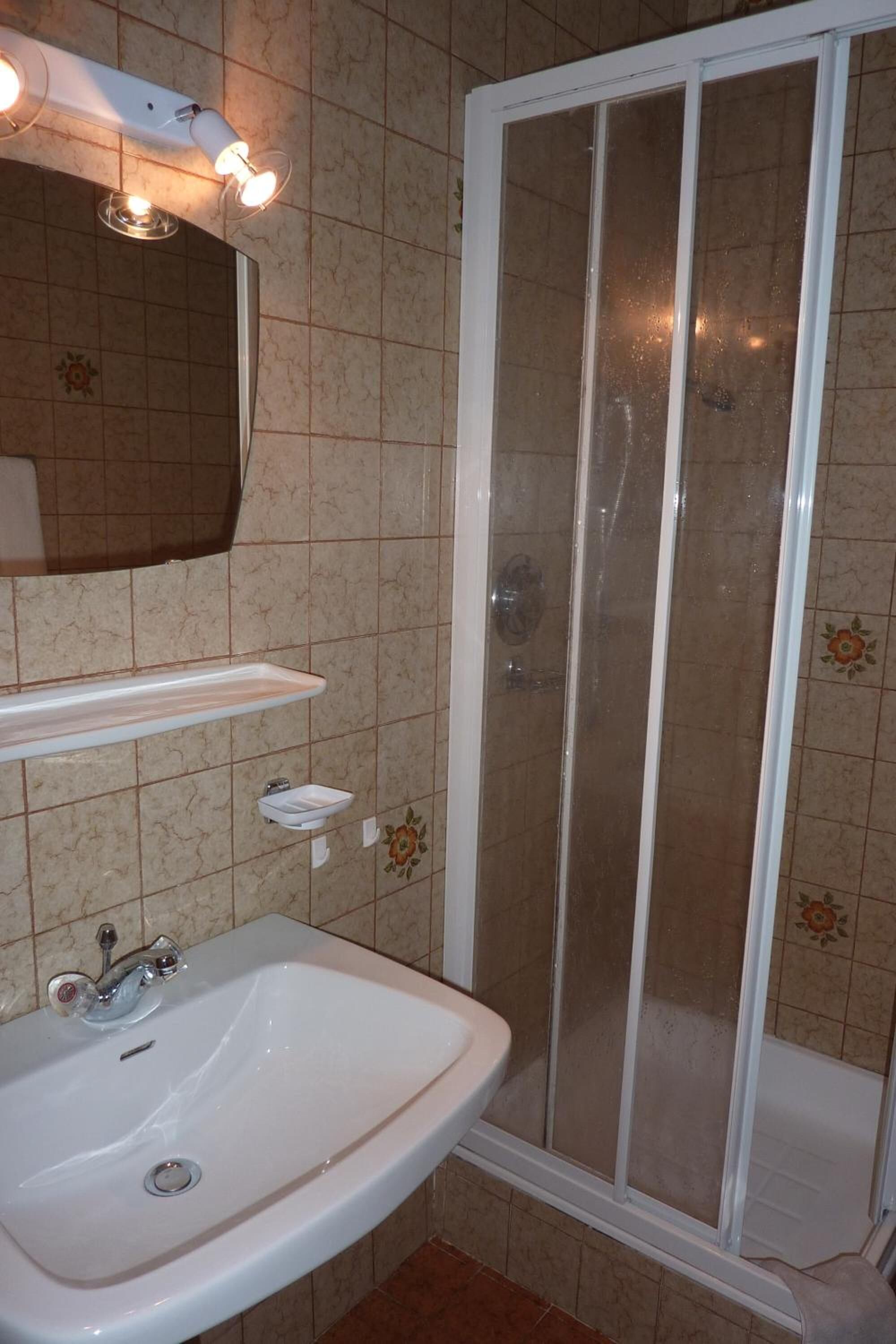 A bathroom with a white sink, a mirror, a shower enclosure, and a tiled wall.