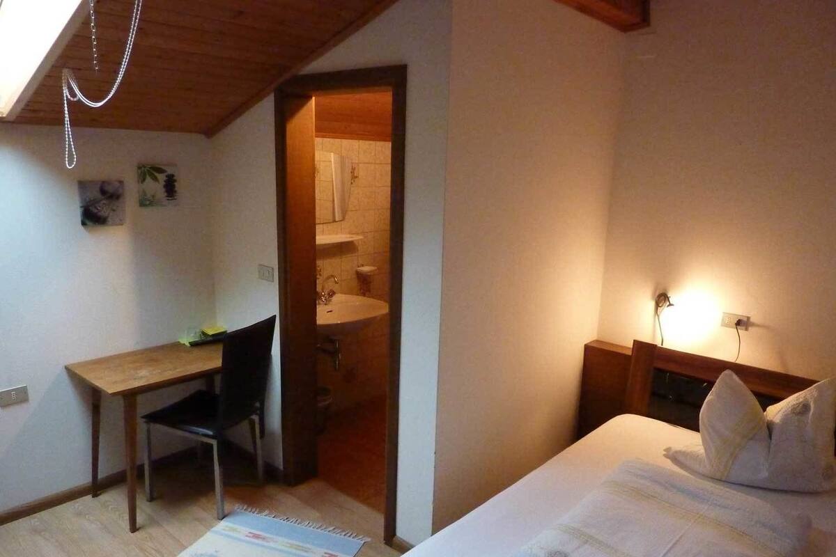 A bedroom with a bed, a desk, and a chair. There is a bathroom visible through an open door.