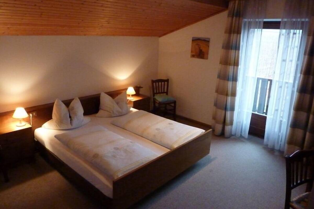 A bedroom with a wooden ceiling, a bed with pillows, two bedside lamps, a chair, and a window with curtains.