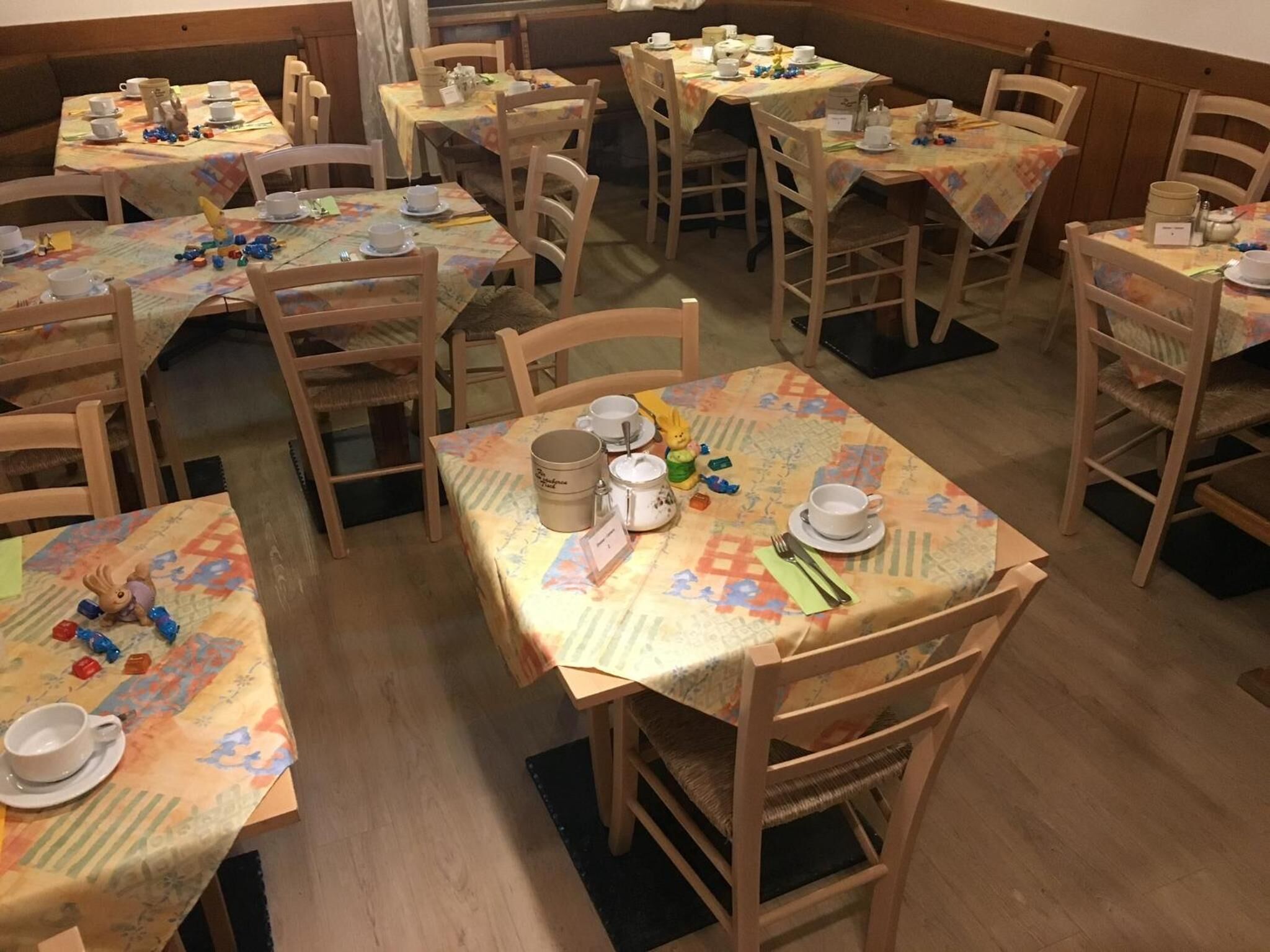 Un comedor con varias mesas, cada una dispuesta con platos blancos, tazas y cubiertos. Las mesas están cubiertas con manteles estampados.