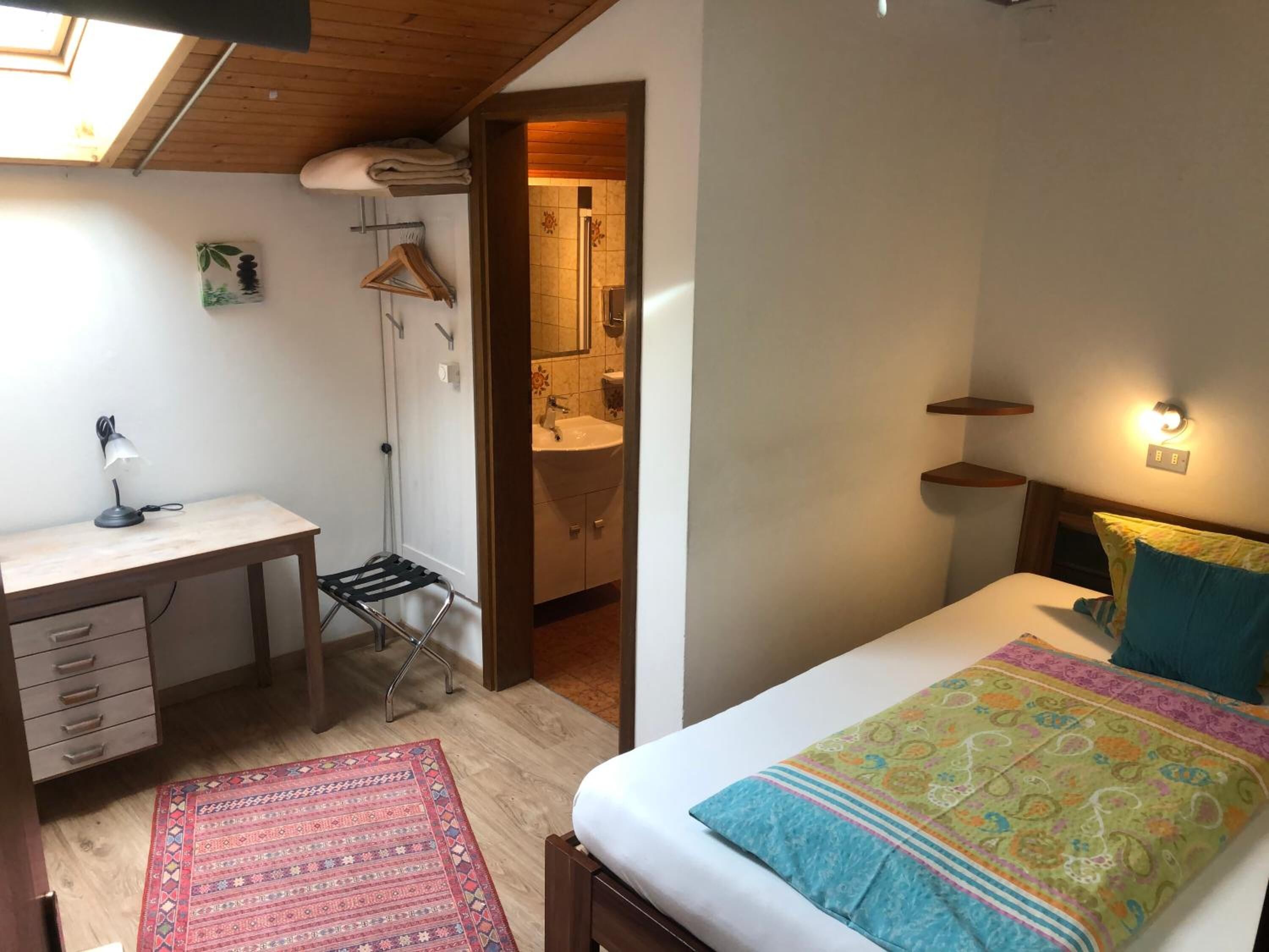 A bedroom with a bed, bedside table, desk, chair, and a small bathroom.
