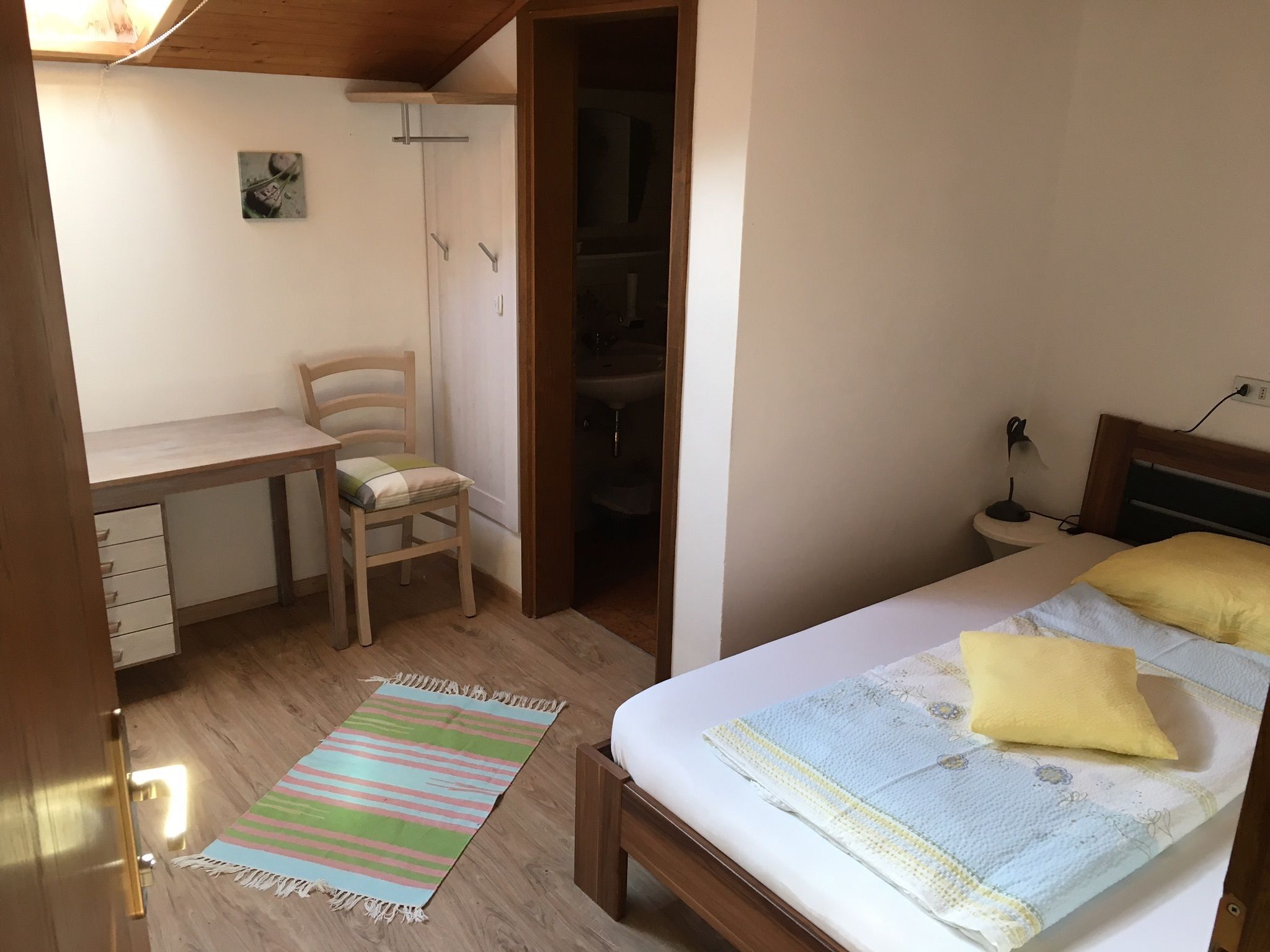 A bedroom with a bed, bedside table, lamp, and a chair. There is a desk with a chair and a small rug on the floor.