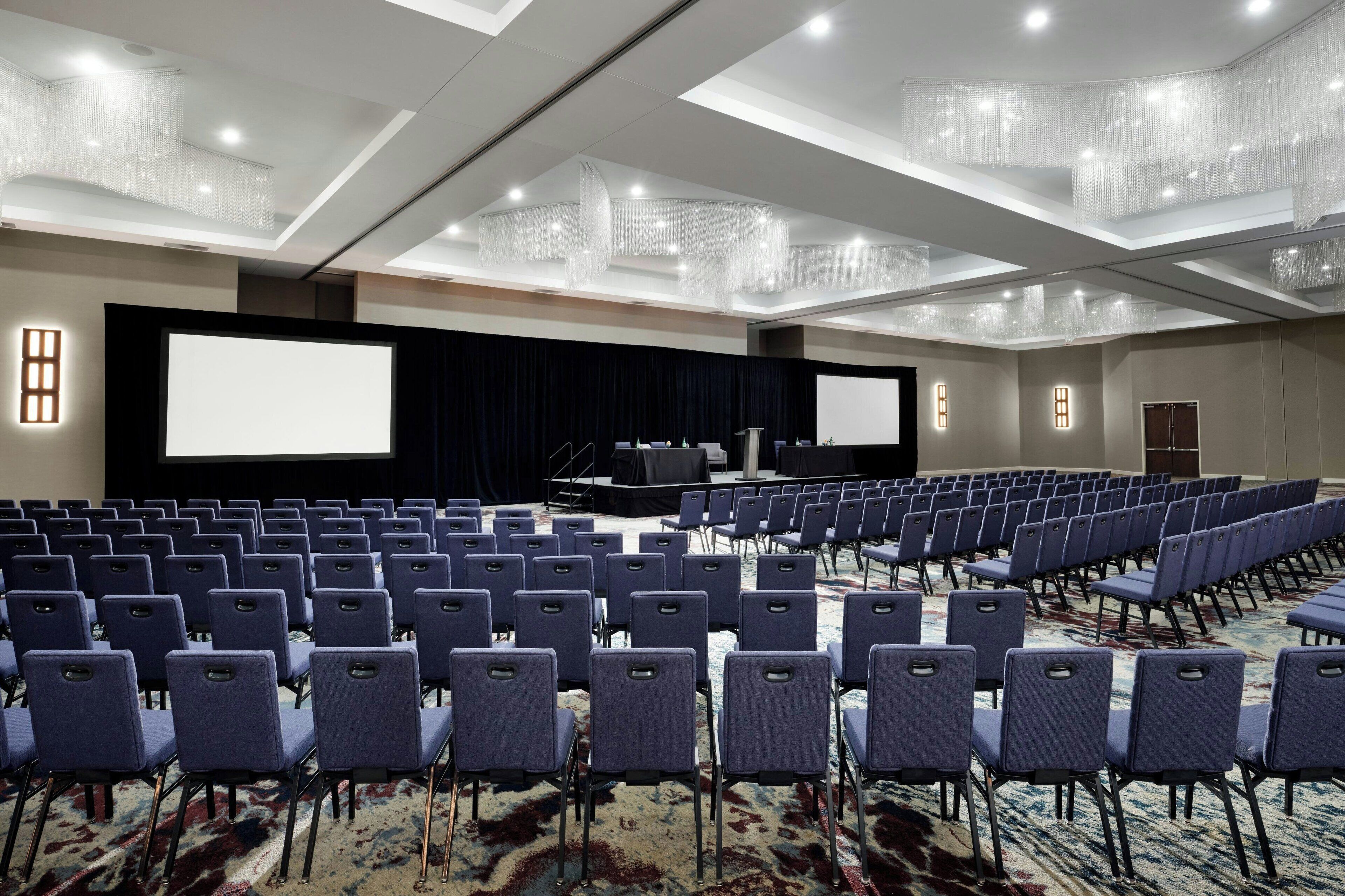 Un salón de conferencias con filas de sillas, una pantalla grande y un escenario.