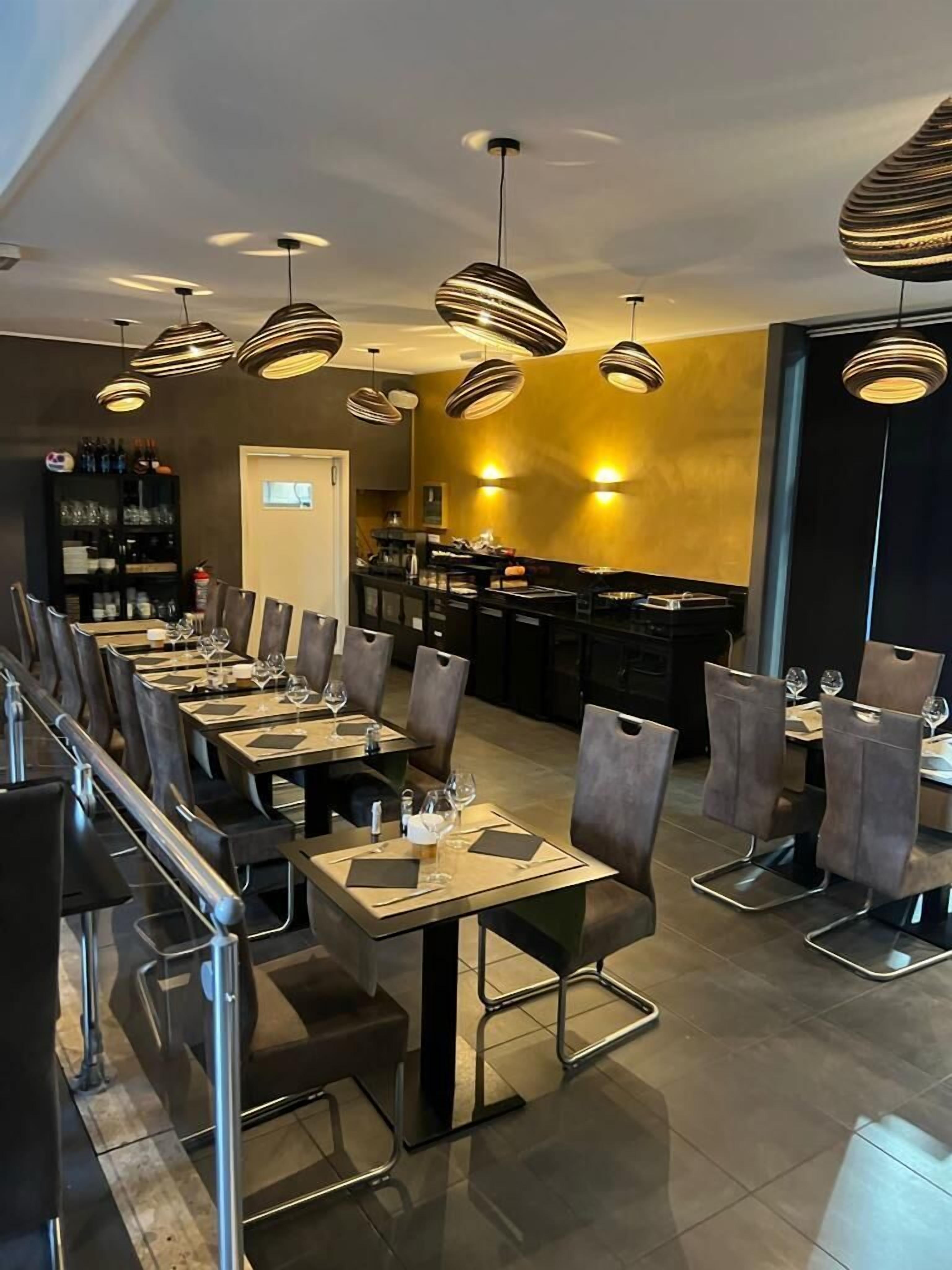 A modern restaurant with a long dining area, black and white striped pendant lights, and a bar area in the background.