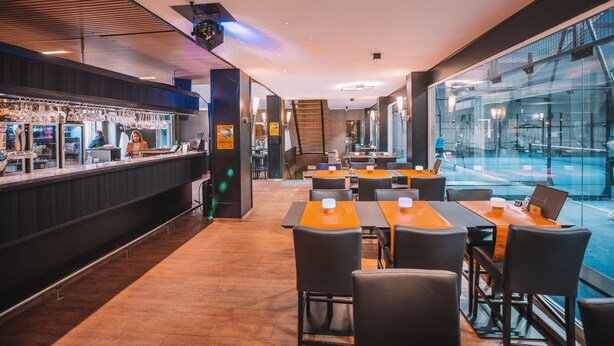 Un ristorante moderno con zona bar, tavoli arancioni e sedie nere, e vista piscina attraverso ampie vetrate.