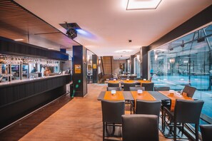 Un restaurante moderno con zona de bar, mesas naranjas y sillas negras, y vistas a una piscina a través de amplios ventanales.