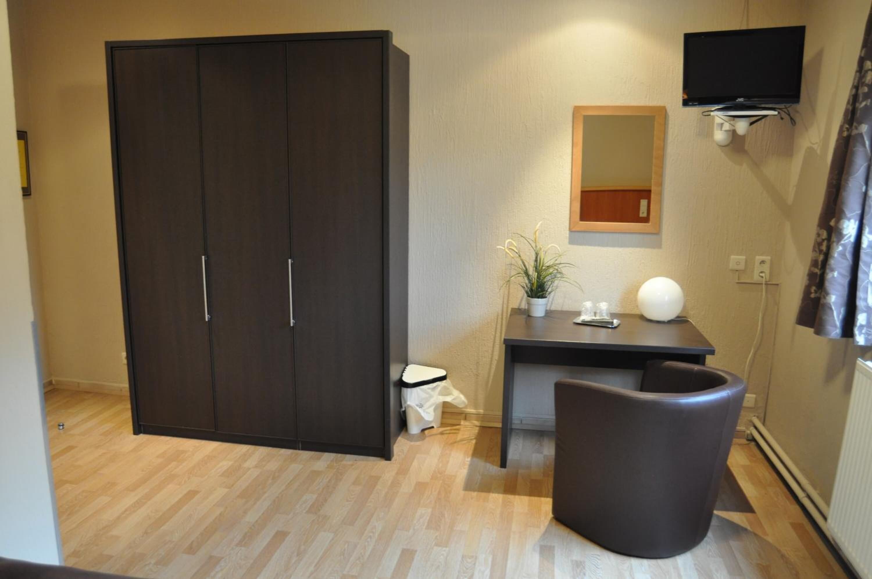 A room with a wardrobe, a desk with a plant, a chair, a small trash bin, and a mounted TV.
