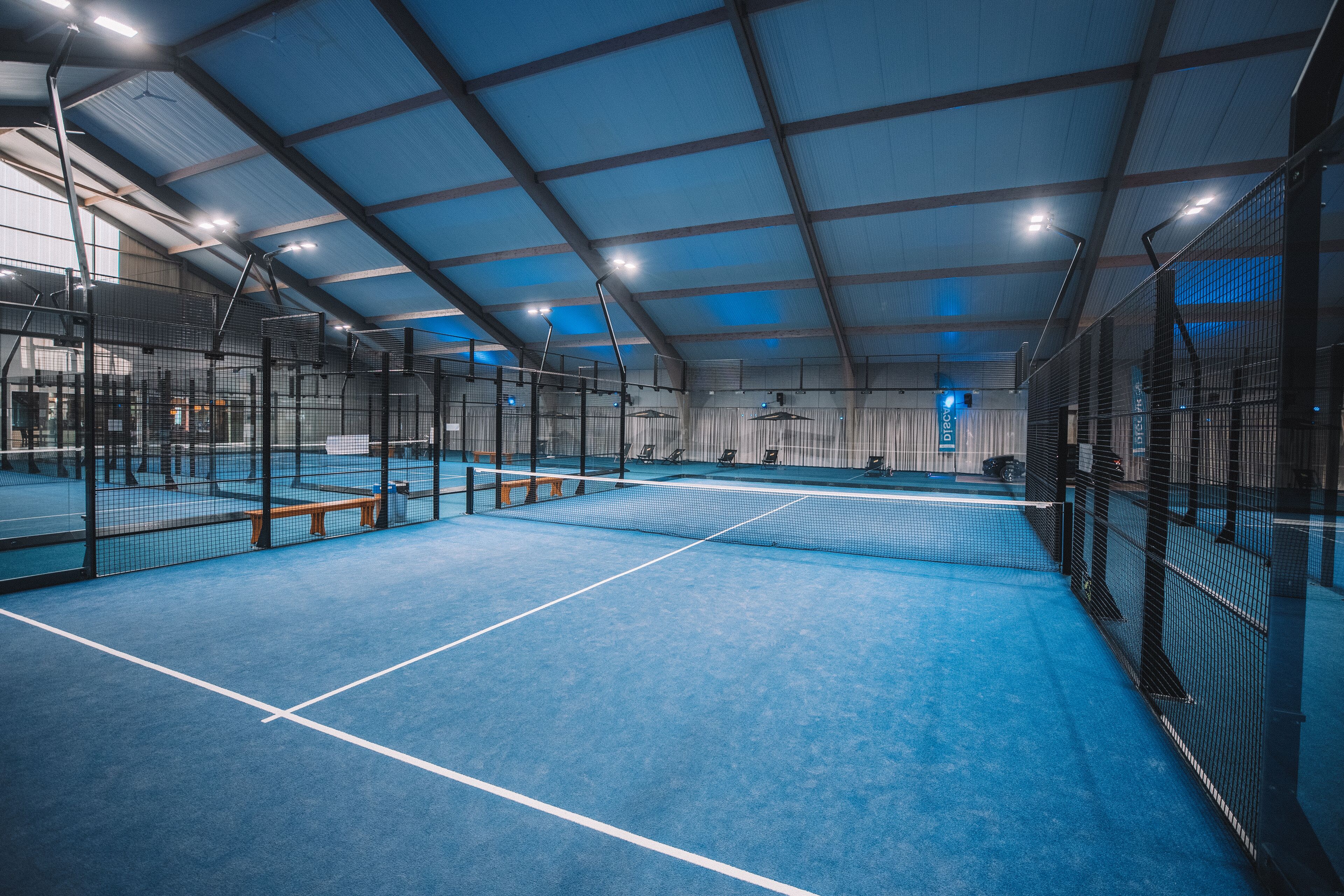 An indoor tennis court with a blue floor, enclosed by metal fencing, and equipped with benches and lighting fixtures.