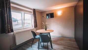 Una habitación con una mesa redonda, dos sillas, una planta en maceta, una ventana con cortinas, un televisor montado en la pared y un radiador.