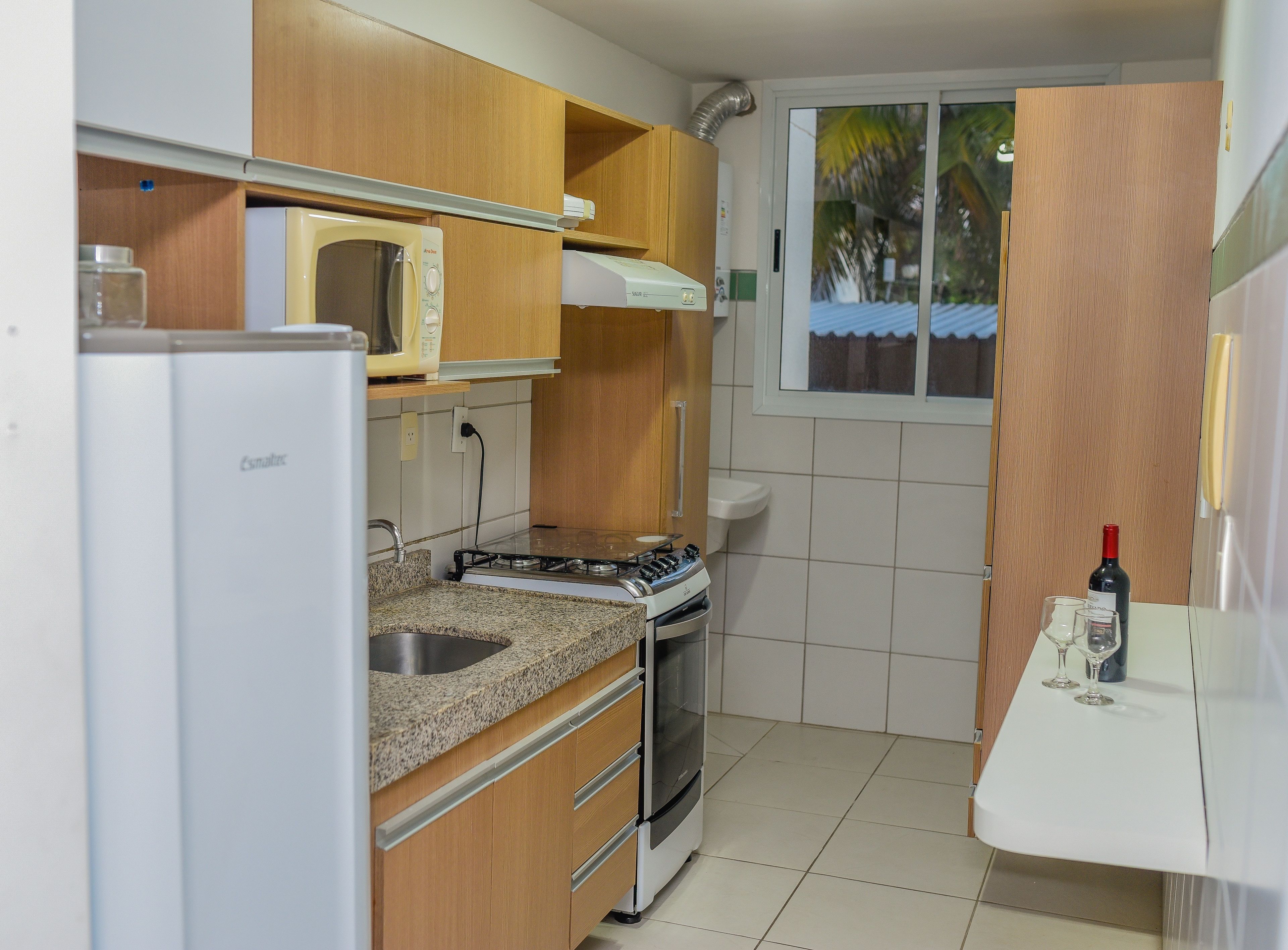 A compact kitchen with wooden cabinets, a microwave, a stove, and a window with a view of palm trees.