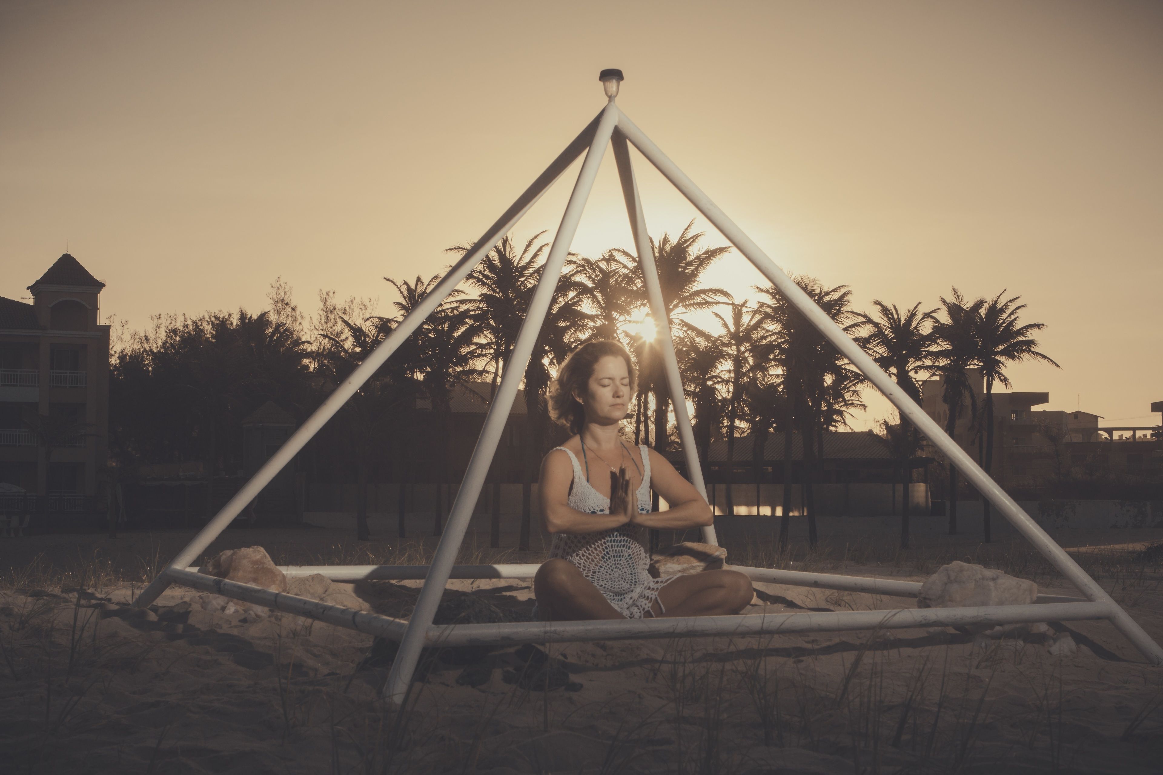 A person sitting in a geometric structure outdoors with palm trees and buildings in the background.