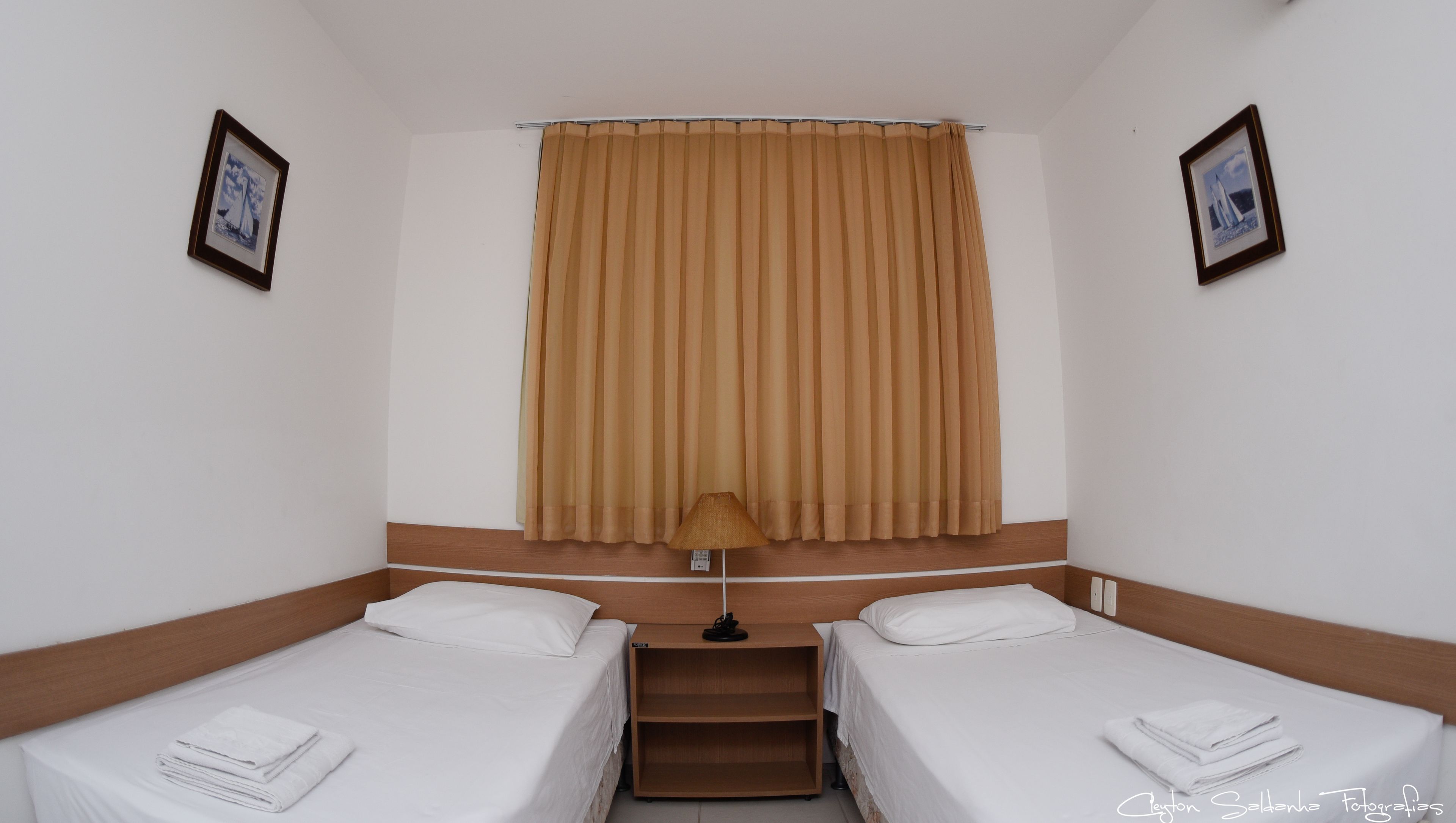 A hotel room with two single beds, a window with beige curtains, a nightstand with a lamp, and two framed pictures on the walls.