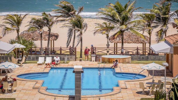 A pool area with a view of the beach, palm trees, and a fenced deck with lounge chairs and umbrellas. - Atlantic Palace Apart-Hotel (Aquiraz)