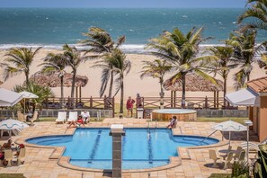 Zona de piscina con vistas a la playa, palmeras y una terraza cercada con sillones de descanso y sombrillas.