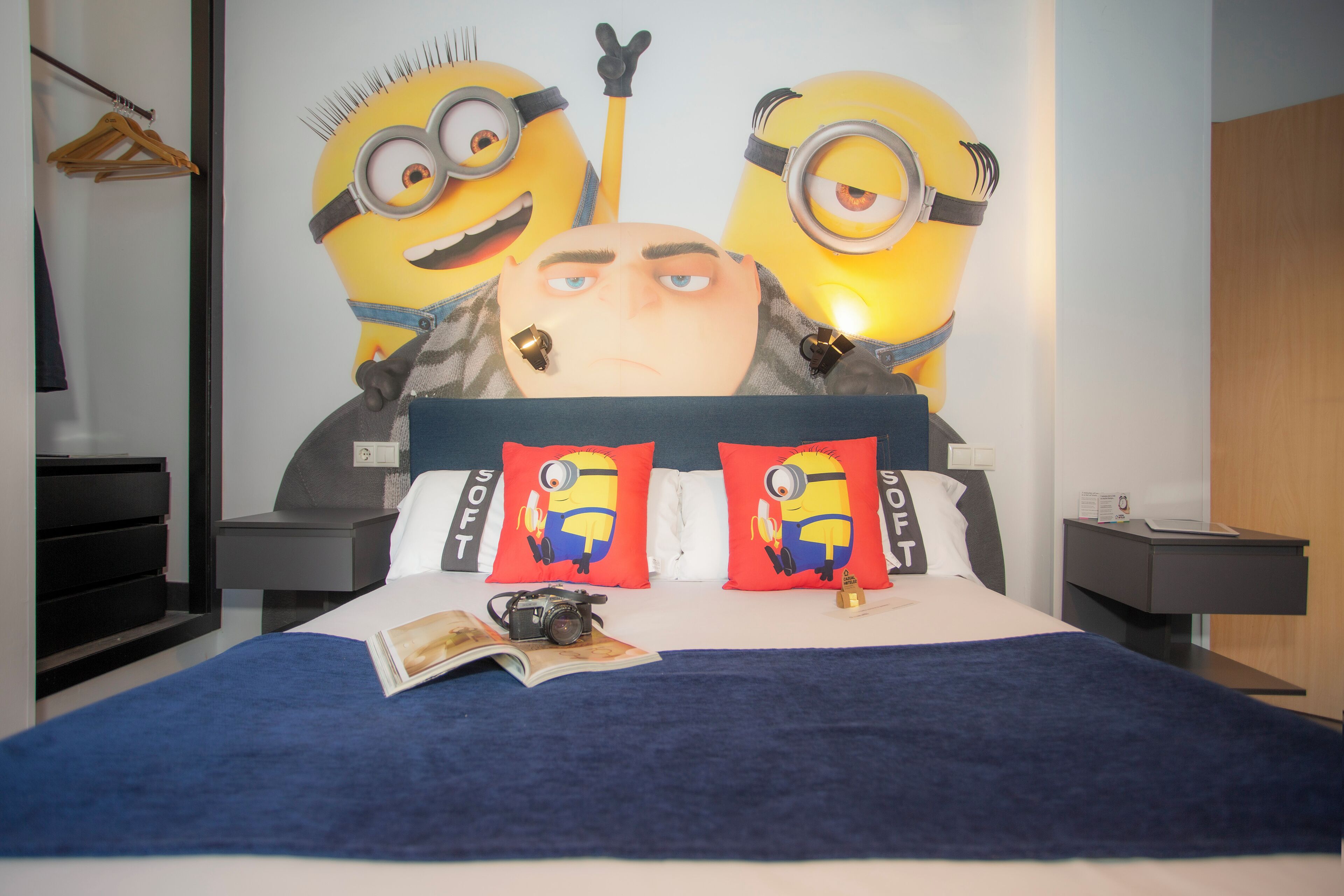 Un llit amb un capçal gran amb temàtica de Minions, dos coixins de Minions, una càmera damunt el llit i una revista.