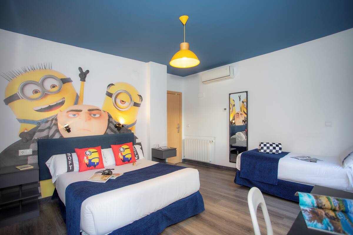 Hotellihuone, jonka seinässä on suuri Minion-aiheinen seinämaalaus, sinivalkoinen vuodevaatteilla varustettu sänky, yöpöytä lampulla ja pieni työpöytä tuolilla.