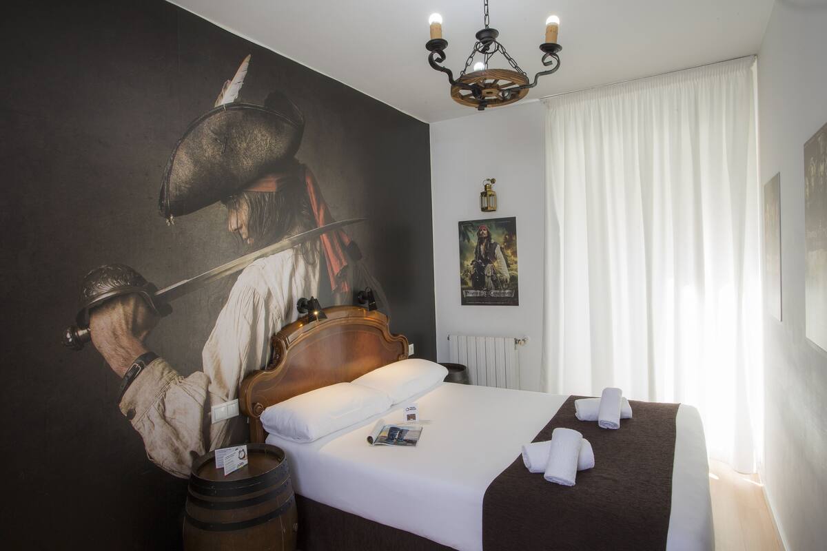 Hotellihuone, jossa on suuri sänky, puinen pääty, valkoiset vuodevaatteet, ruskea heittopeitto ja seinällä kuva historiallisesta henkilöstä.
