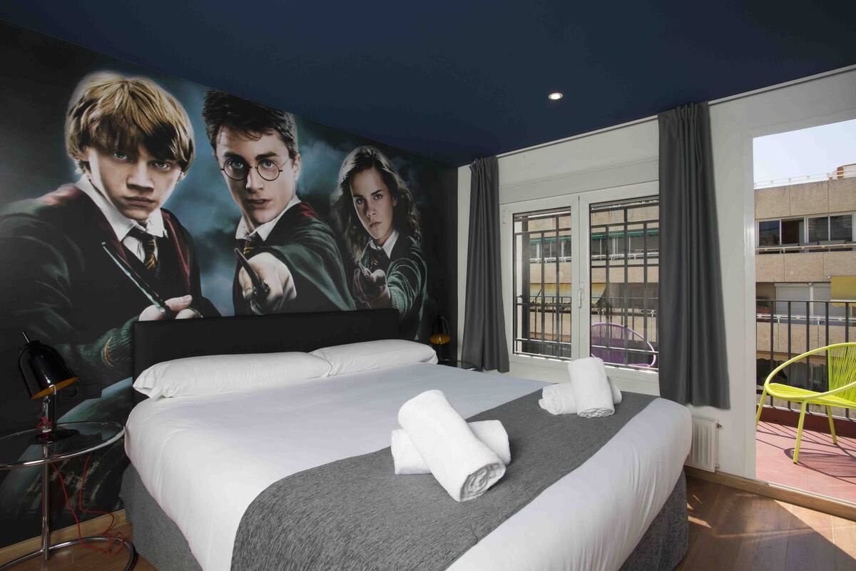 Hotellihuone, jonka seinällä on suuri Harry Potter -hahmojen juliste, siististi pedattu sänky valkoisilla lakanoilla ja harmaalla päiväpeitteellä, pieni pyöreä pöytä lampun kanssa ja näkymä rakennukseen ikkunasta.