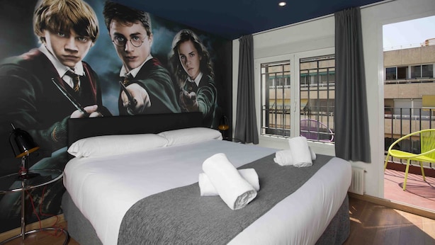 Habitación de hotel con un cartel grande de personajes de Harry Potter, una cama bien hecha con sábanas blancas y un plaid gris, una pequeña mesa redonda con una lámpara, y vista a un edificio por la ventana.