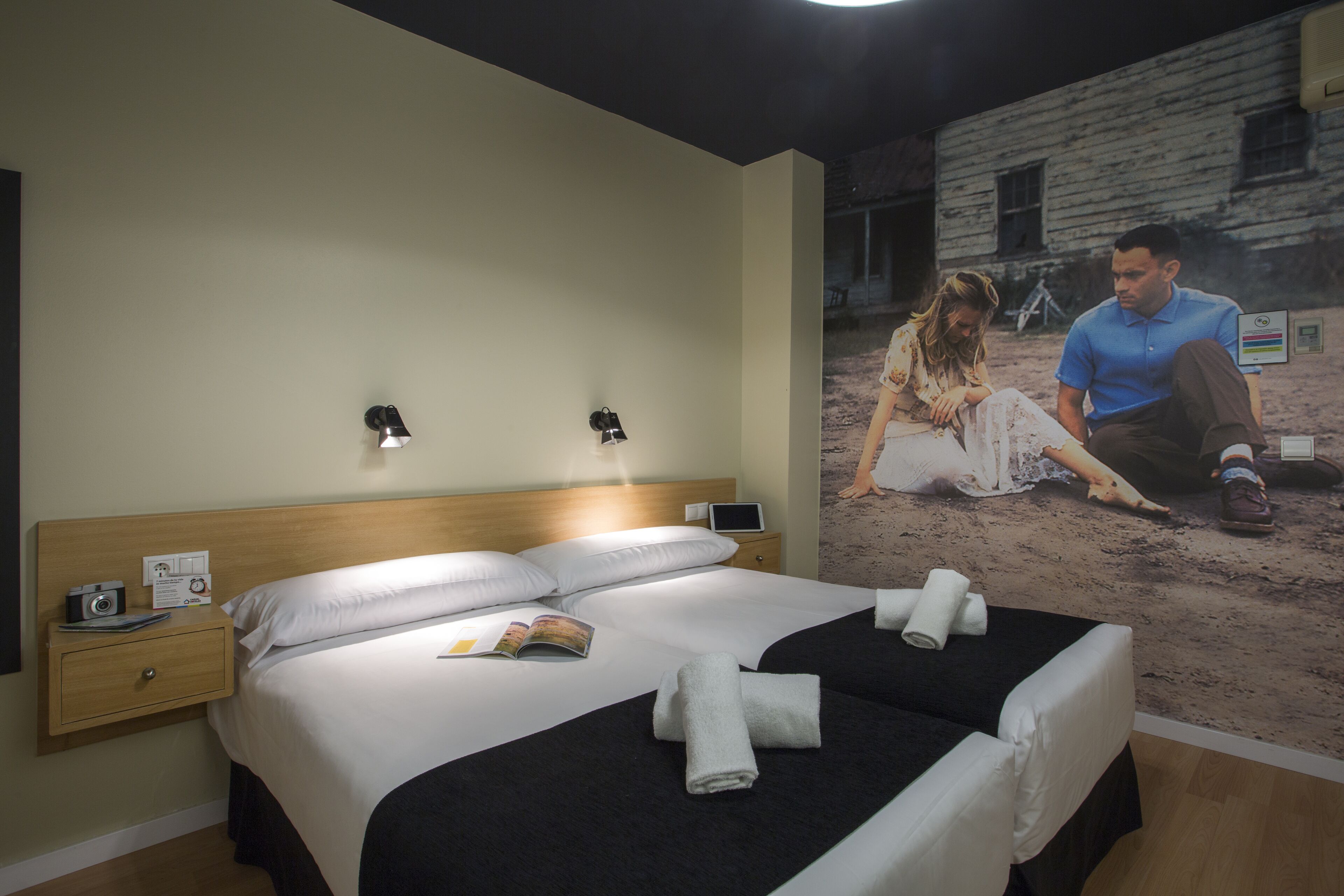 Ein Hotelzimmer mit einem großen Bett, Nachttischen, einem Beistelltischchen mit einer Lampe und einem Poster an der Wand.