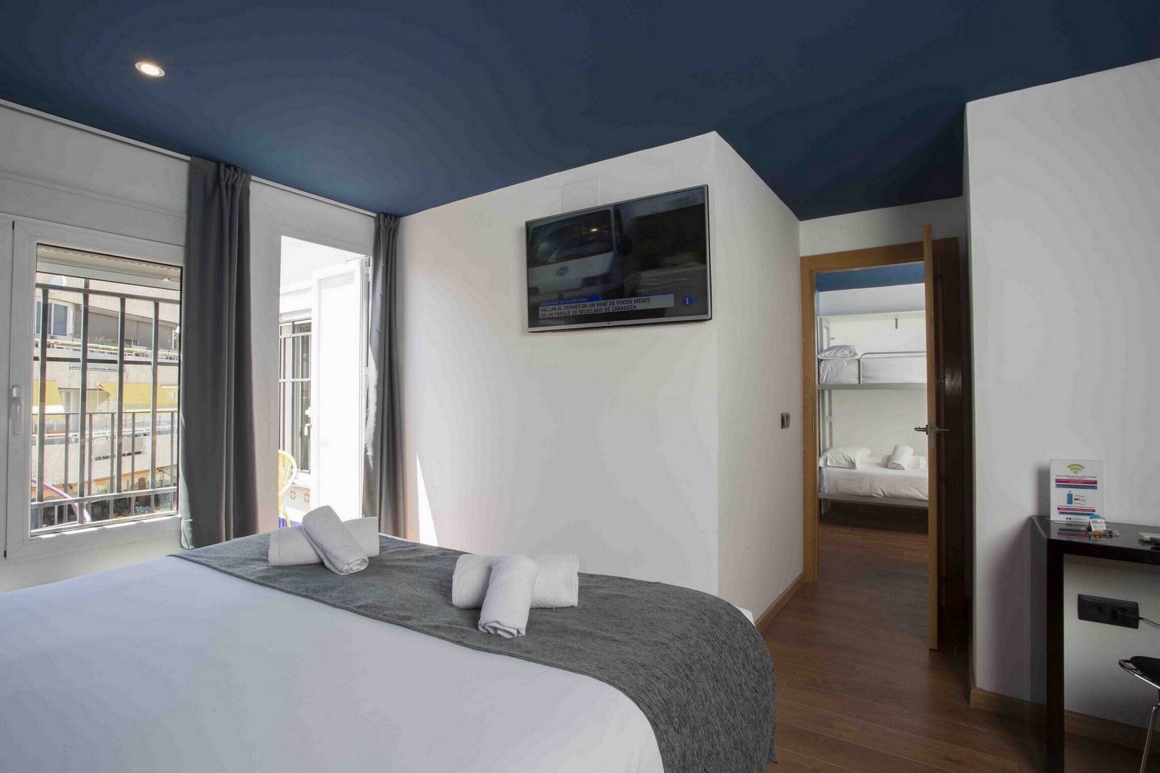 A hotel room with a bed, a TV mounted on the wall, a desk, and a view of the outside through a window.
