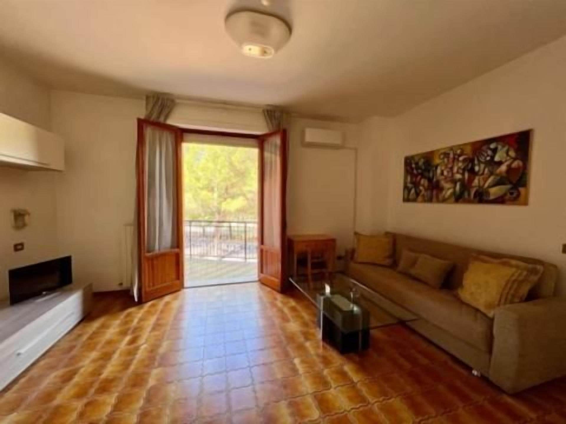 Una sala de estar con sofá, televisor y chimenea. Tiene un balcón con vistas a árboles.