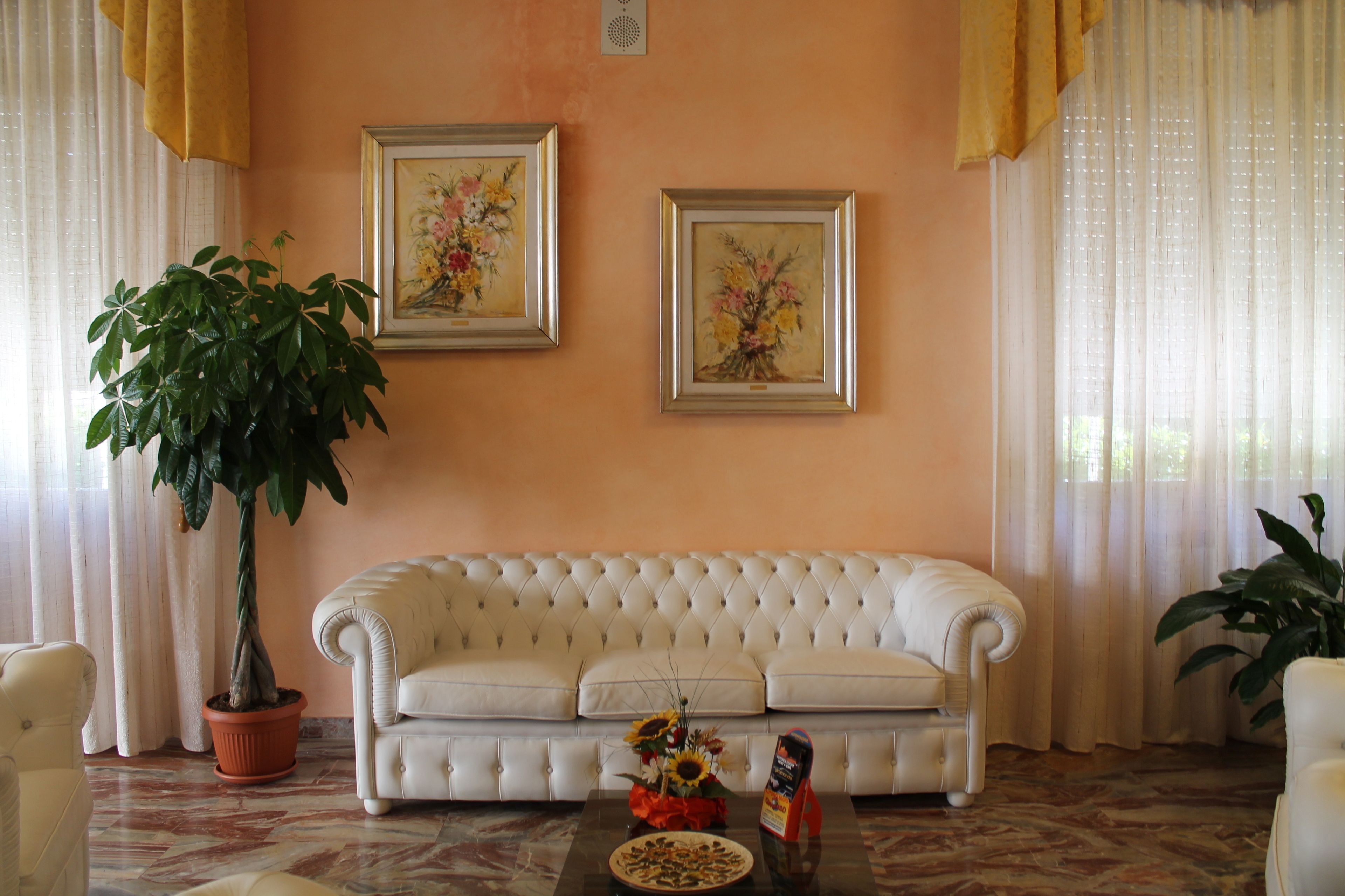 A living room with a white tufted sofa, two framed floral paintings on the wall, a potted plant, and a coffee table with a vase of flowers.
