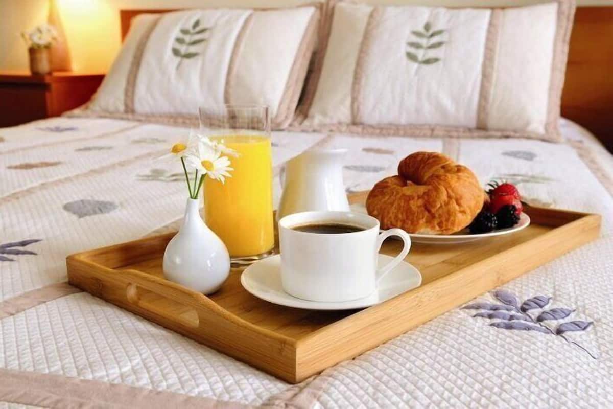 A breakfast tray on a bed with a cup of coffee, a glass of orange juice, a croissant, and a small vase with a flower.