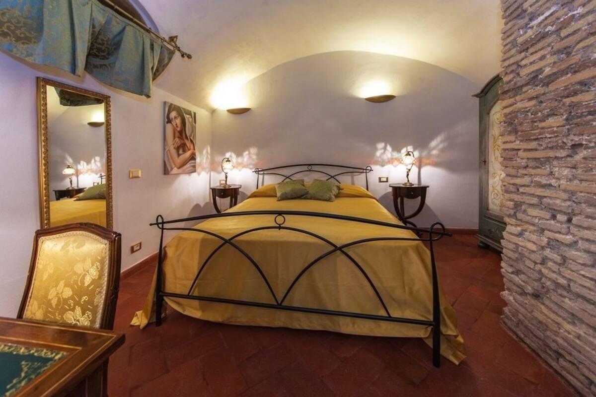 A bedroom with a bed, a mirror, a painting, and a stone wall.
