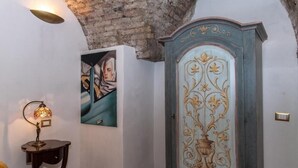 A room with a rustic brick ceiling, a wall-mounted painting, a decorative cabinet with a floral design, and a table lamp. - Rifugio Degli Artisti (Rome)
