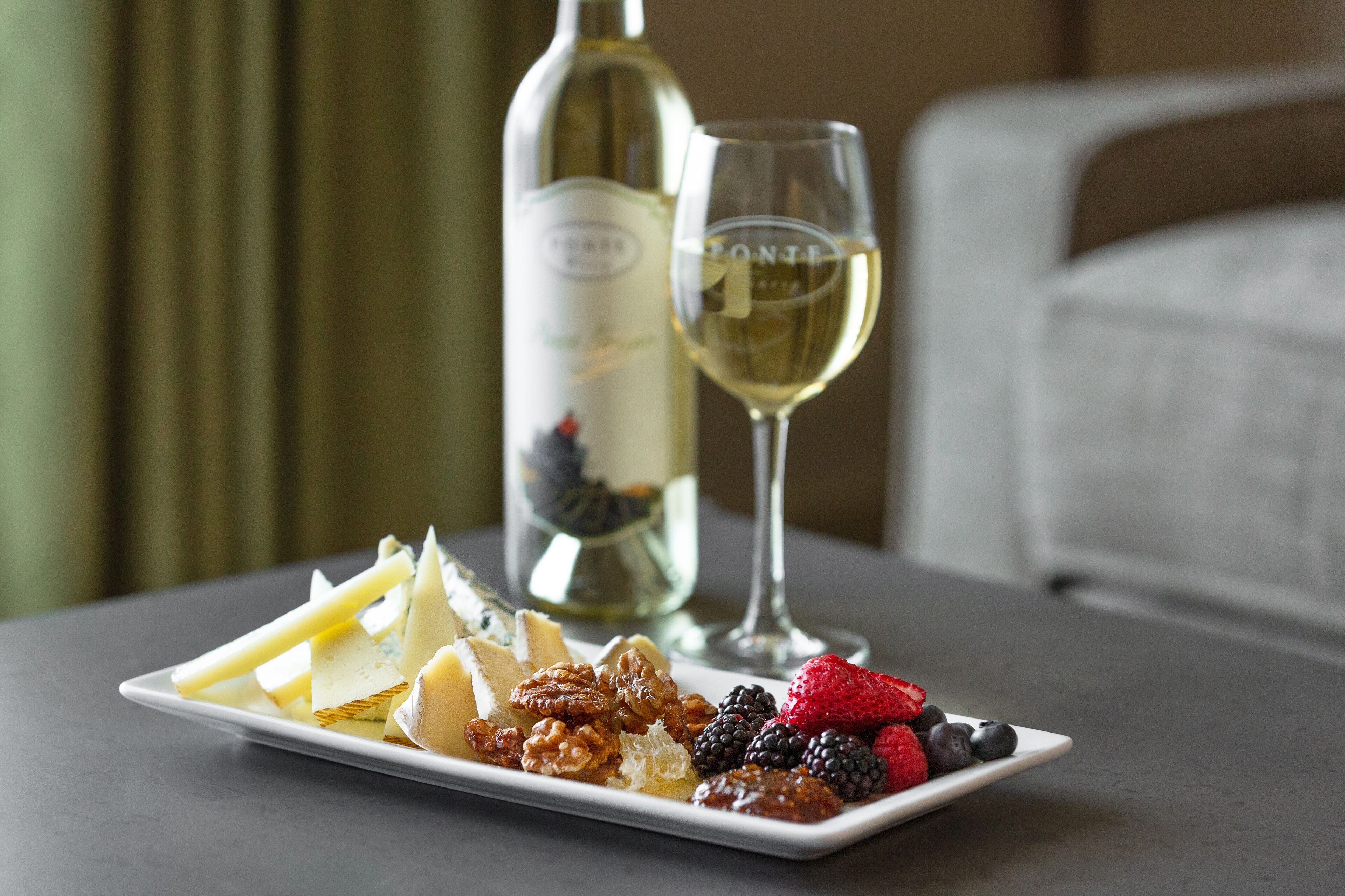 A plate of assorted cheeses, nuts, and berries on a table with a bottle of white wine and a glass.