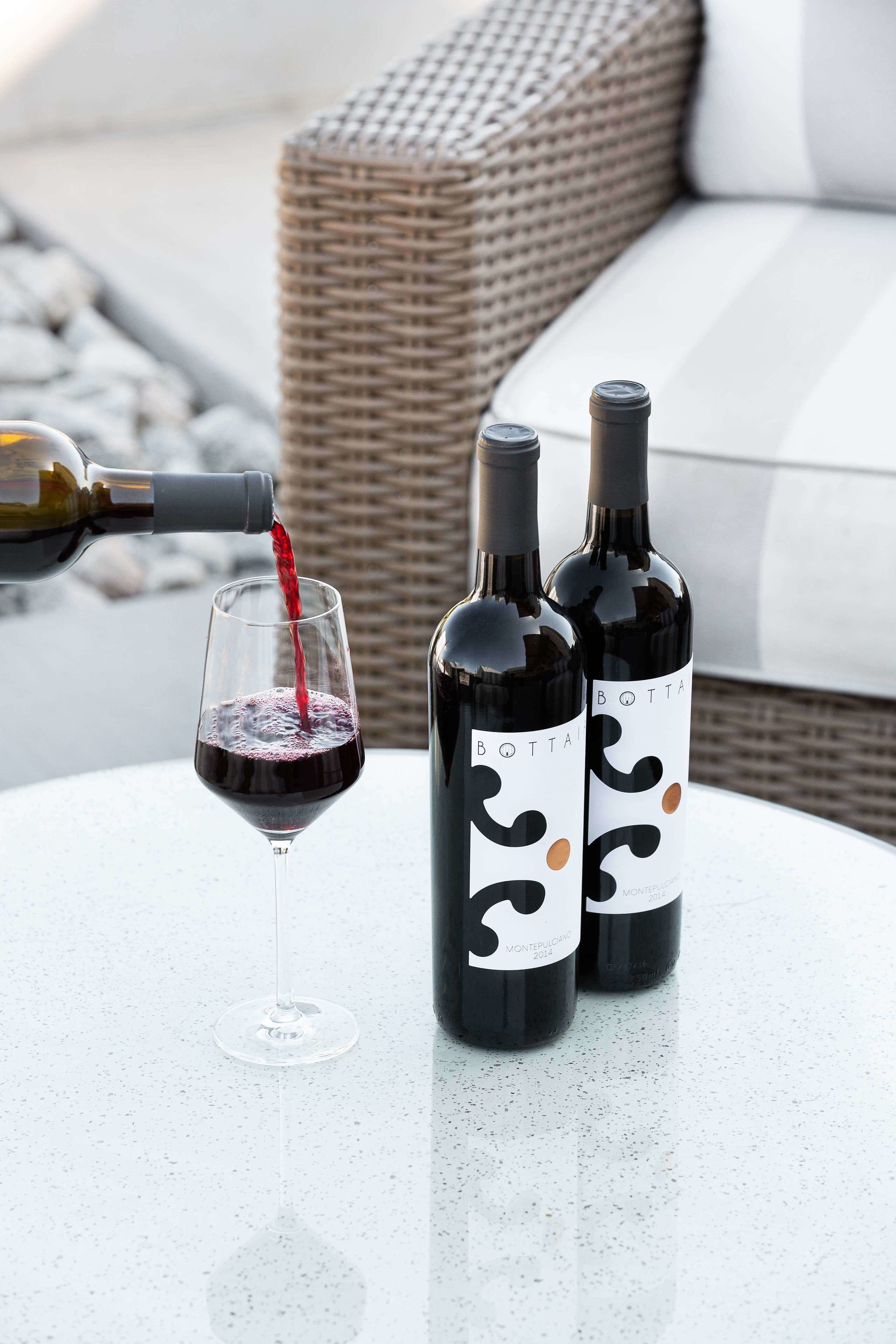 Two wine bottles and a glass on a table with a wicker chair in the background.