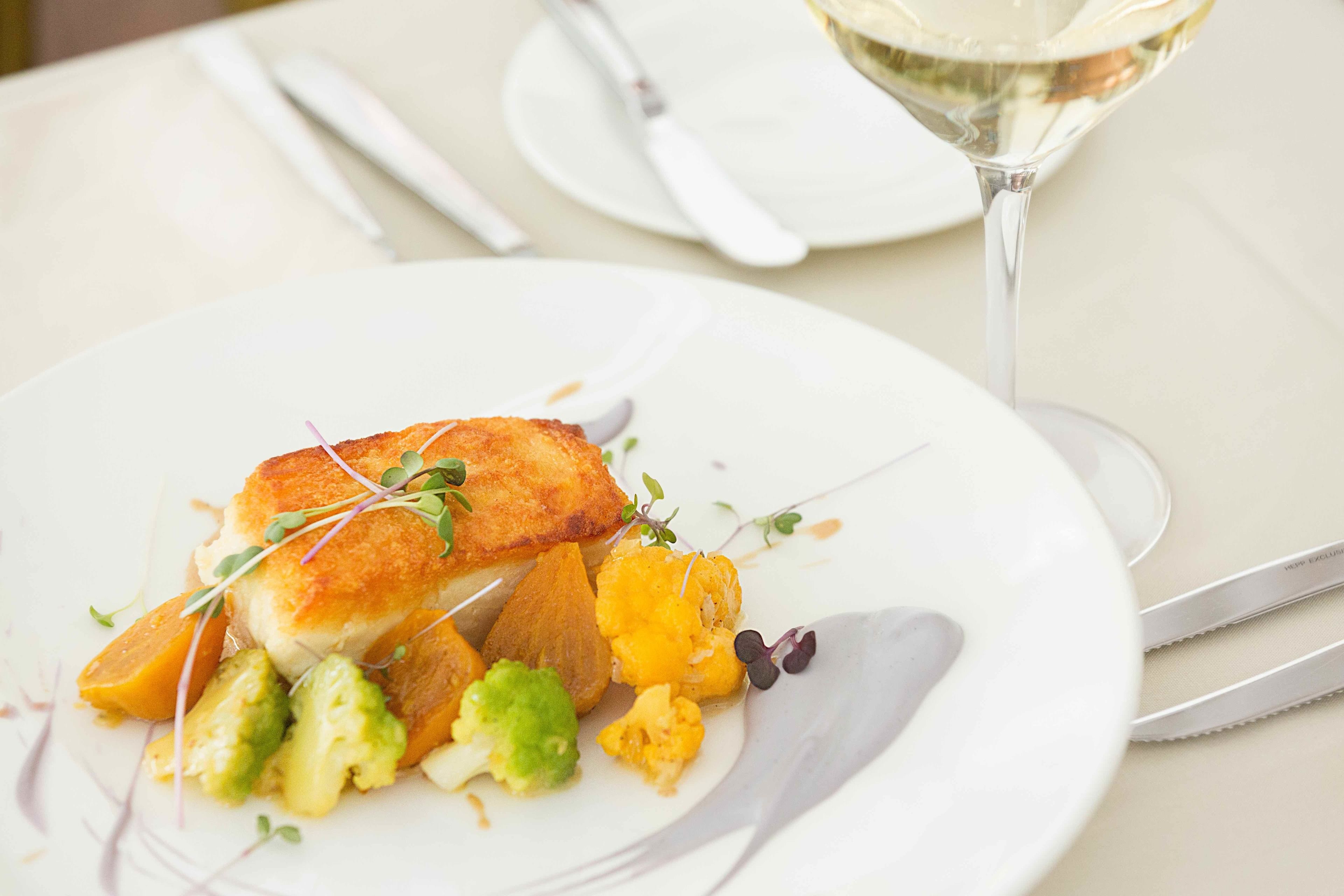 A plate of food with a fish, vegetables, and a glass of white wine on a table.