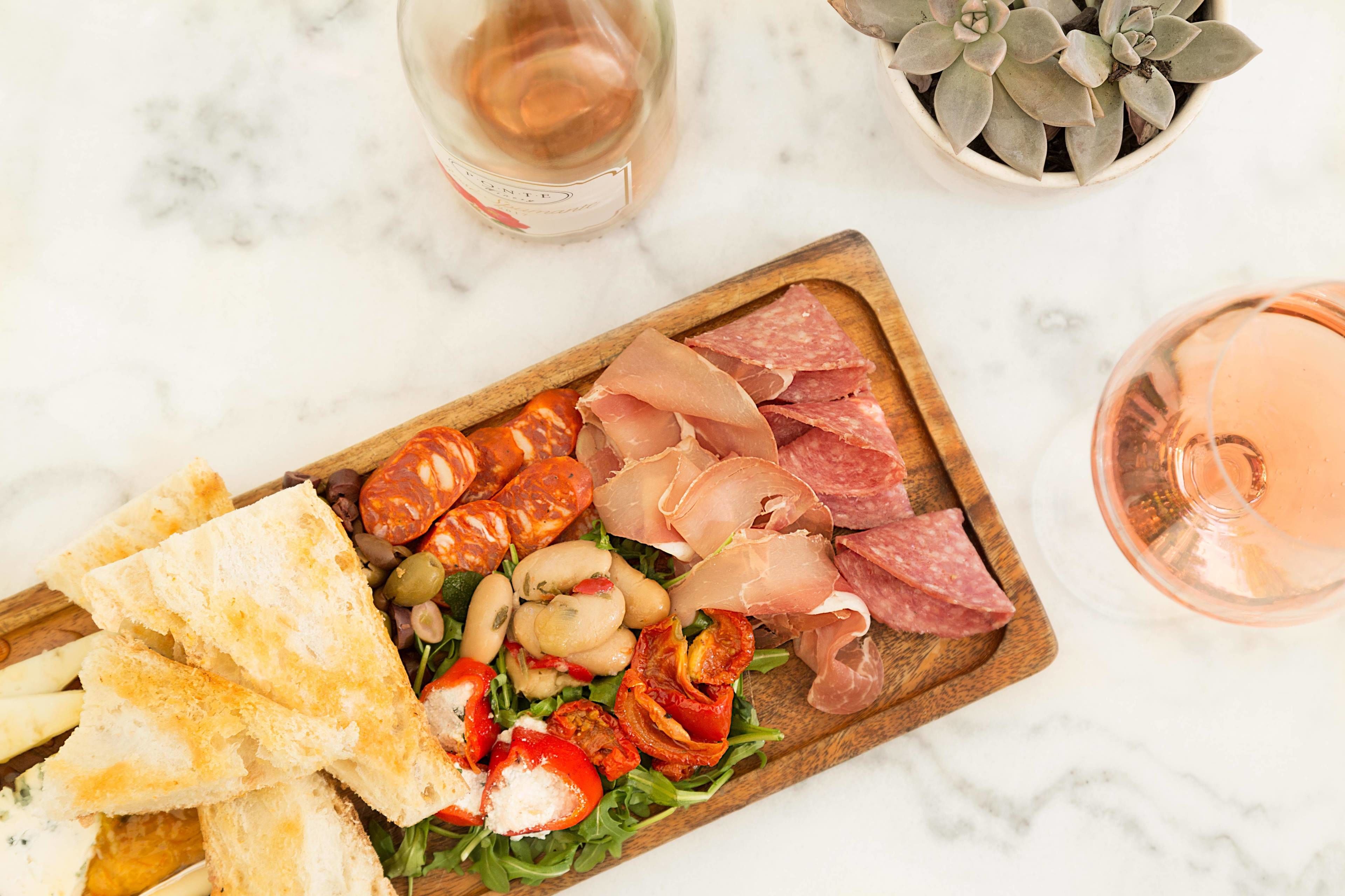 A charcuterie board with various meats, cheeses, and vegetables on a wooden board, accompanied by a glass of rosé wine and a potted plant.