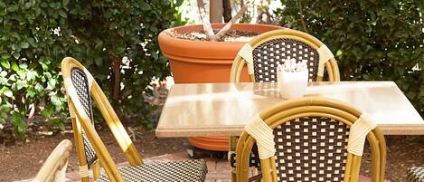 An outdoor patio with wicker chairs and a table, surrounded by potted plants and a hedge.