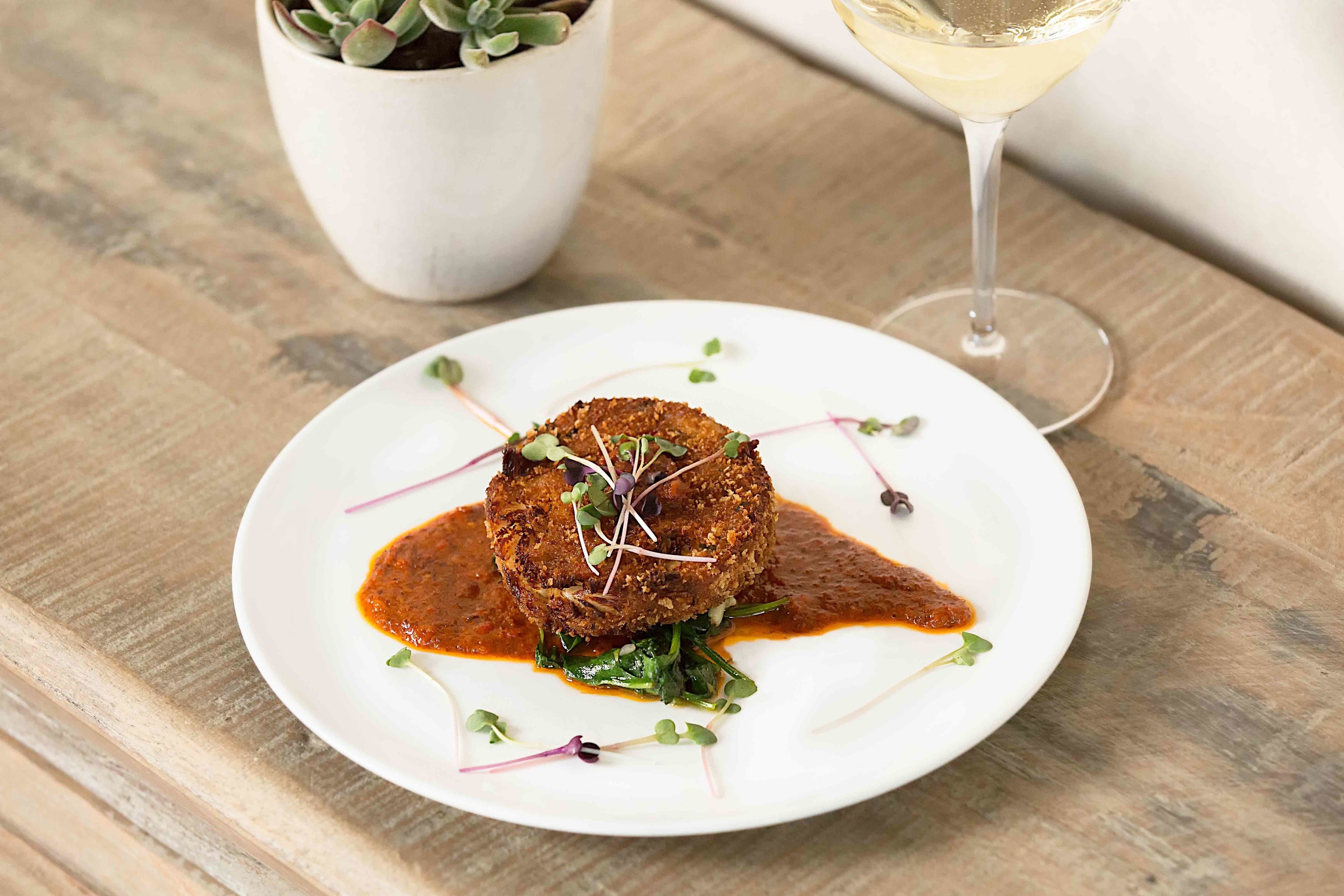 A plate of food with a sauce and greens, a glass of white wine, and a potted plant in the background.