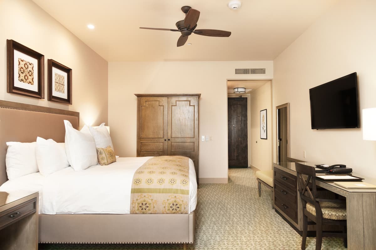 Habitación de hotel con cama, escritorio, silla, ventilador de techo, armario y televisor en la pared.