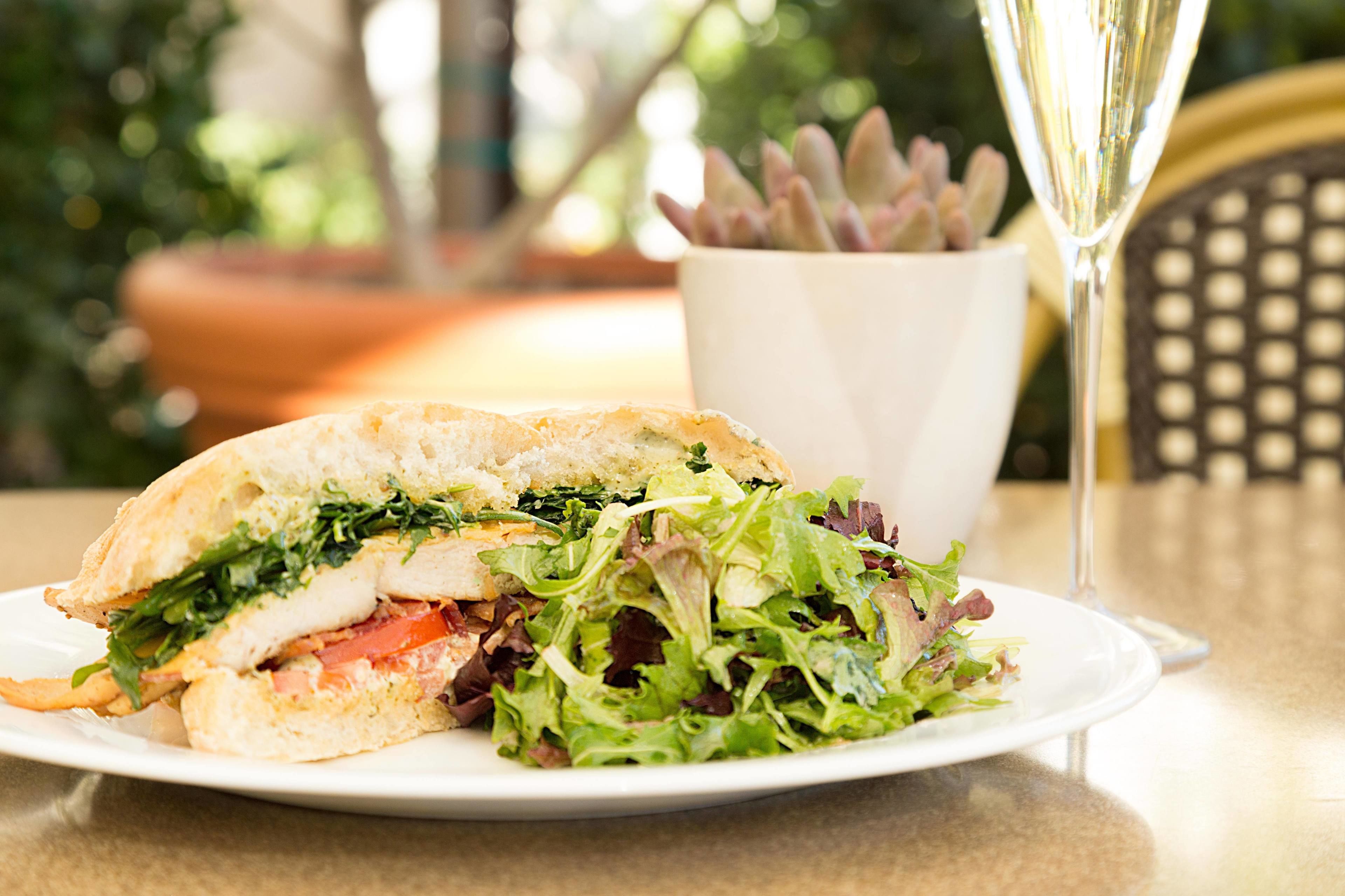 A sandwich on a plate with a glass of champagne and a potted plant in the background.