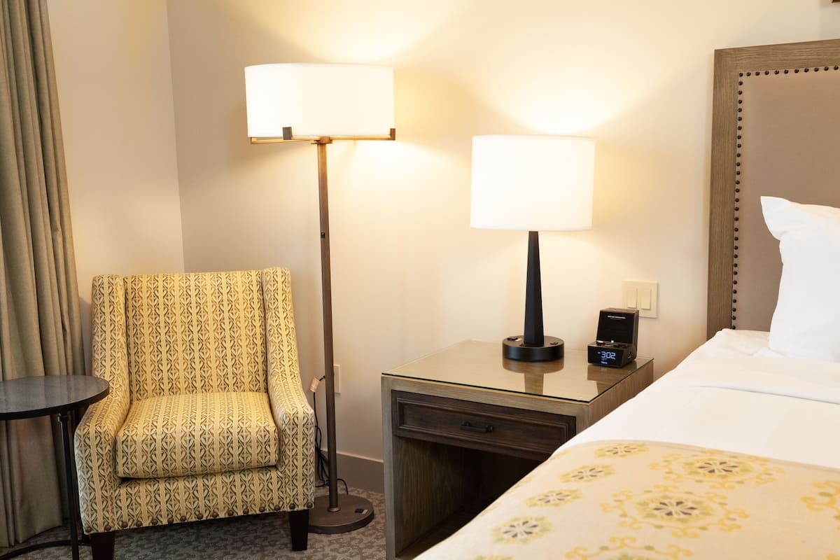 Una habitación de hotel con un sillón estampado, una mesita de noche con una lámpara, un reloj y una cama con edredón estampado.