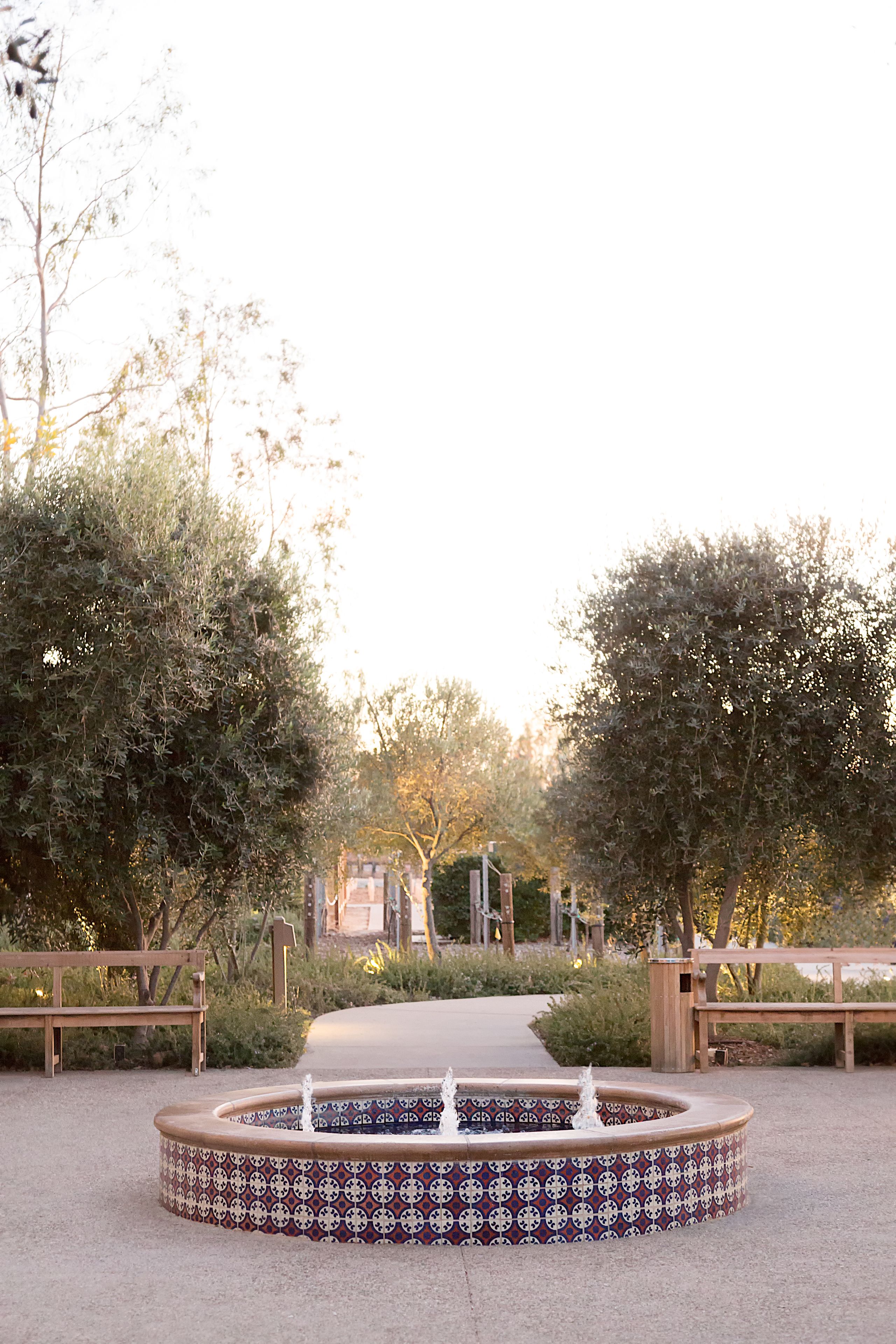 Ein runder Brunnen mit einem gemusterten Design, umgeben von Bänken und Bäumen in einer Parkanlage.