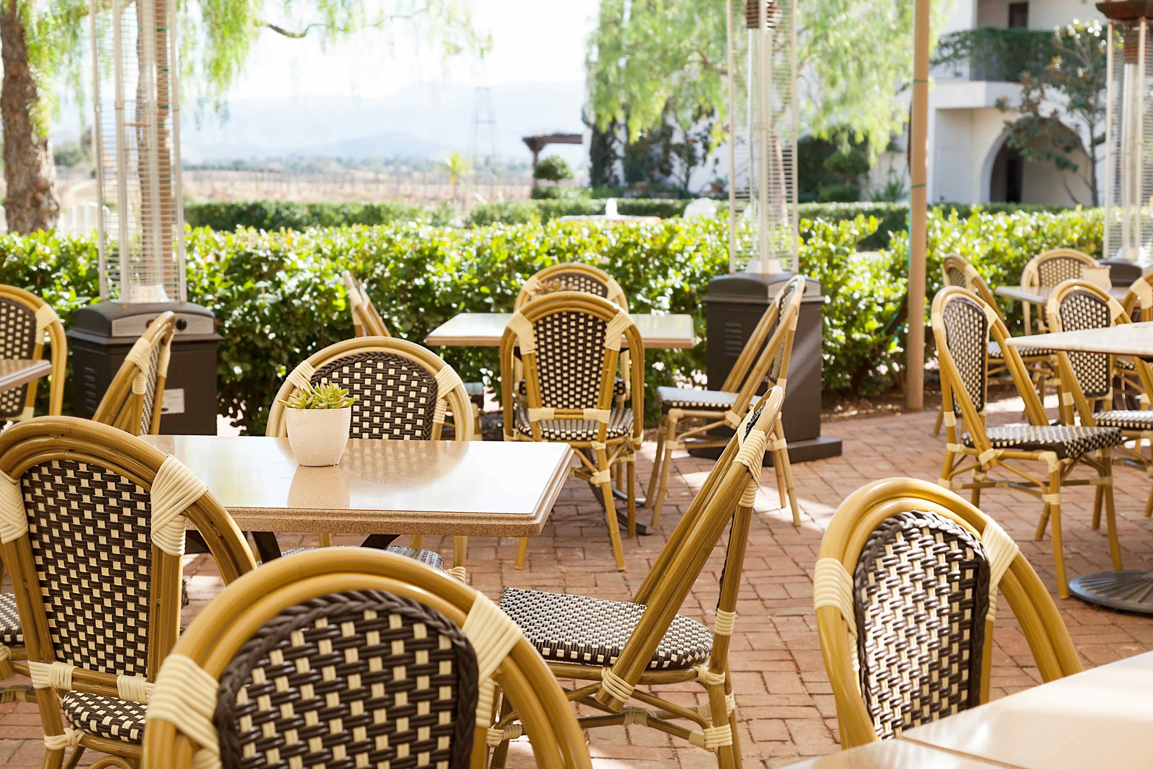 An outdoor seating area with wicker chairs and tables, surrounded by greenery and a view of the landscape.