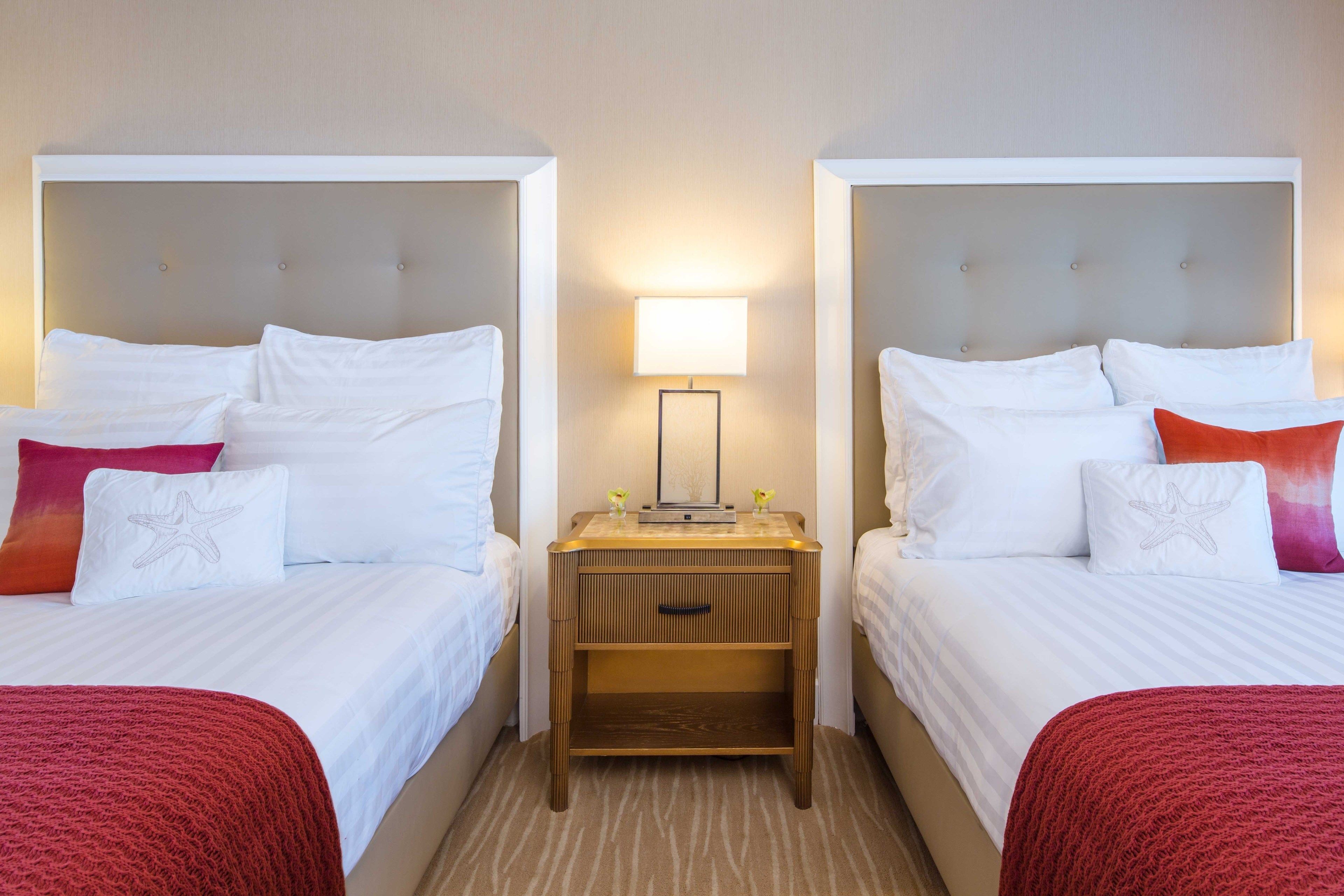 Ein Hotelzimmer mit zwei Betten, jeweils mit weißer Bettwäsche und roten Akzenten, einem Nachttisch mit Lampe sowie einer kleinen Ablagefläche.