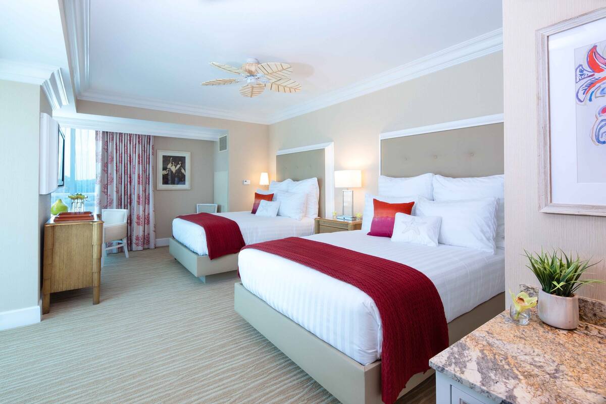 A hotel room with two beds, a desk, a chair, a ceiling fan, and a potted plant.