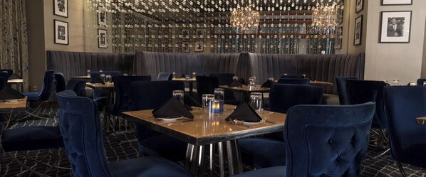 A restaurant with dark blue upholstered chairs, tables with black napkins, and a chandelier.