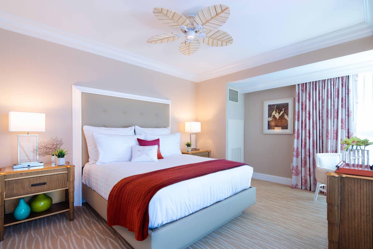 A hotel room with a large bed, two bedside tables, a nightstand, a ceiling fan, a window with curtains, and a framed picture on the wall.